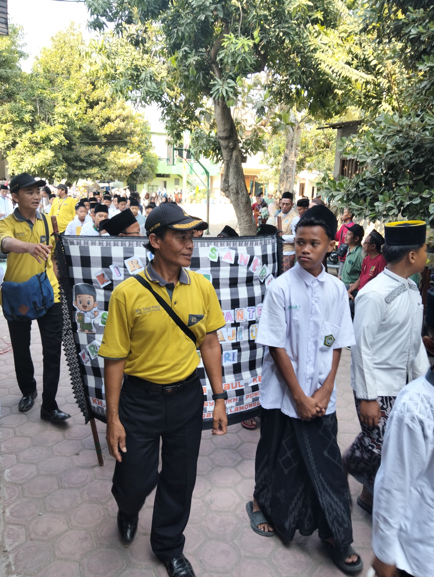 In Kranji, Indonesia, a group of people, likely students and staff, are walking outdoors on a paved path. The foreground features two men in yellow polo shirts and black pants. The man on the left is wearing a blue bag across his chest and a black baseball cap. The man on the right, also in a black cap, is carrying a checkered banner with cartoon characters and text. Behind them, a line of young people, mostly wearing white shirts and black or white caps, are walking. Some of the young people are wearing traditional Islamic attire. In the background, there are trees and buildings, suggesting an outdoor school or community event. The lighting indicates it is daytime, possibly morning or afternoon, with sunlight filtering through the trees. The overall scene depicts a procession or a walk, possibly for a school event or religious observance.
