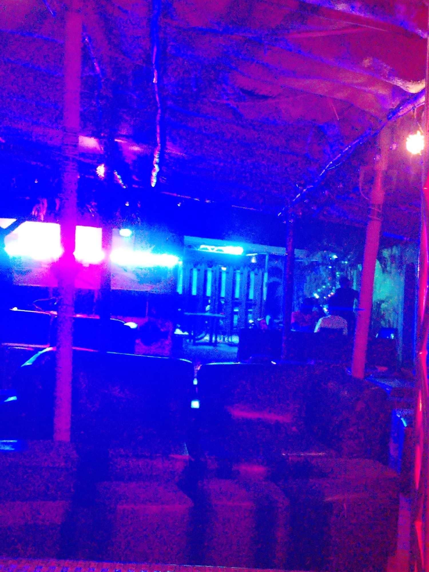 The image depicts a dimly lit indoor or semi-enclosed space, likely a bar, lounge, or a casual entertainment venue, dominated by strong artificial lighting. The primary colors in the scene are vibrant blues and magenta/pink, creating a dynamic and atmospheric ambiance characteristic of a nighttime setting.
Several individuals are present, mostly appearing as dark silhouettes against the glowing lights. In the mid-ground right, two people are seated at a table or booth, possibly socializing. Another person stands further back towards the center, while additional patrons are faintly visible in the background.
The setting incorporates rustic structural elements, including prominent wooden poles or beams that support a textured ceiling, which appears to be made of thatch or similar natural materials. The foreground and mid-ground feature dark, cushioned seating arrangements, possibly booths or benches. Bright, glowing light sources, resembling neon or LED strips, emit intense blue and pink light across the space. A single warm, yellowish light bulb is visible in the upper right corner, contrasting with the cooler tones. In the background left, a slatted wooden structure, possibly a partition or wall, is also illuminated by the colorful lights.
The scene suggests a relaxed social gathering or leisure activity. No visible text is present in the image.