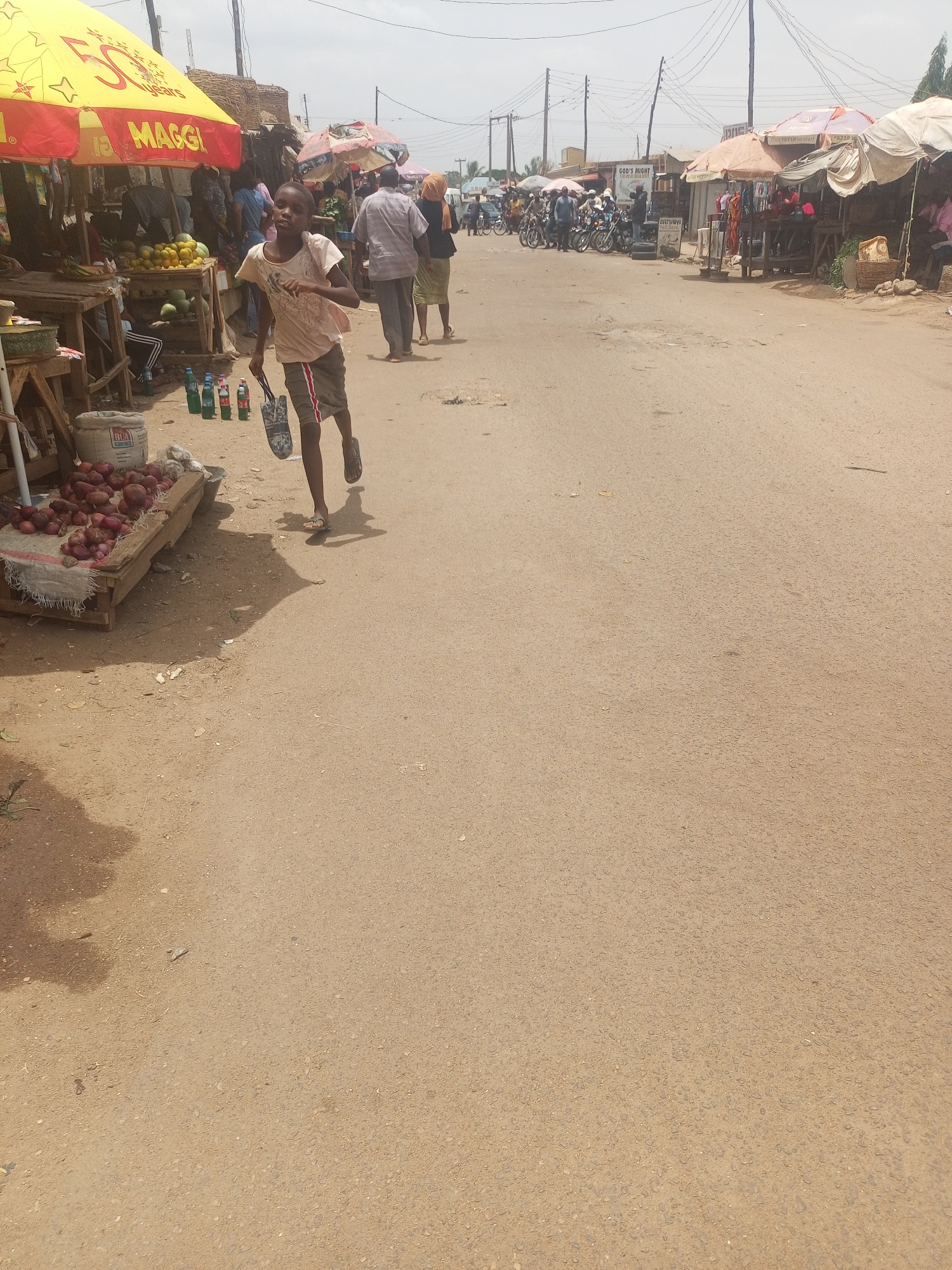 A child is running down a dusty road in Sabon Tasha, Nigeria. The child is wearing a light-colored shirt and dark shorts, and is carrying a bag. The road is lined with stalls covered by umbrellas and yellow Maggi tarps. People stand around the stalls. Some people are standing around motorcycles further down the road. There are onions for sale in a crate on a table to the left of the child. There are also bottled drinks. The sky is overcast.