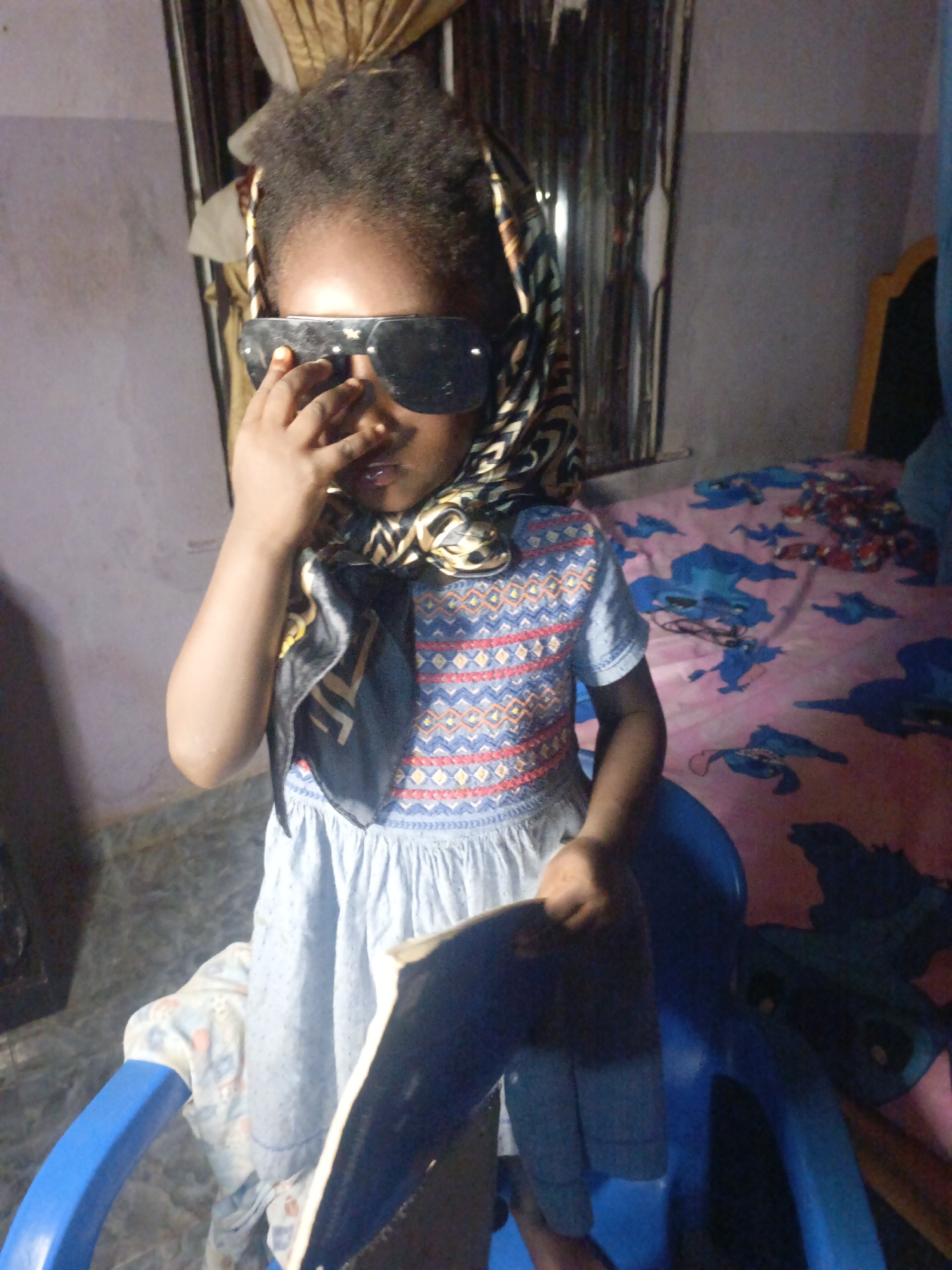 A young child stands in an indoor space, presumably in the city of Akwanga, Nigeria. The child is wearing a blue dress with a patterned yoke, oversized sunglasses, and a patterned scarf draped over their head. The child holds a book or magazine in one hand and touches the sunglasses with the other. The background shows a bed with a patterned sheet and curtains. The child stands behind a blue plastic chair with a white patterned cloth beneath her.