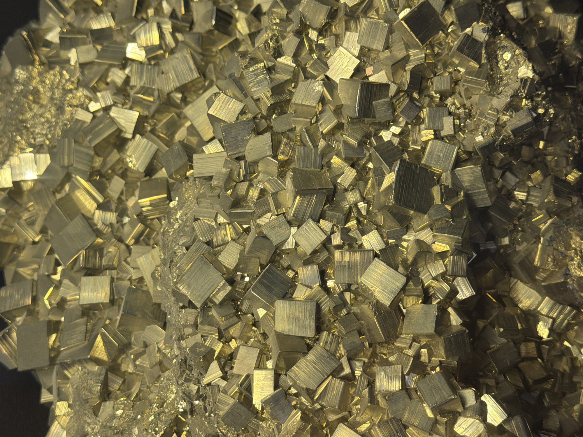 The image displays a close-up, overhead view of a large pile of pyrite crystals, also known as "fool's gold." The crystals are primarily cubic in shape and exhibit a metallic, brass-yellow luster with varying degrees of reflectivity due to the lighting. Some of the crystals show striations on their faces. The overall texture of the pile is rough and uneven, with many of the crystals clustered together. The background is dark and out of focus, with a hint of blackness at the top left. The scene is an abstract composition focusing on the geometric forms and metallic sheen of the mineral specimens. There are no people, actions, or interactions depicted. The image does not contain any information that would indicate a specific location within Fort Lauderdale, United States of America.