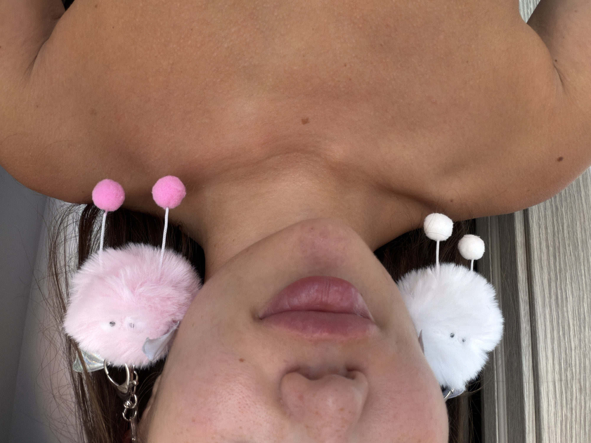 A close-up, chest-up shot shows a person wearing large, fluffy pom-pom earrings. The earrings are white and pink, with smaller pom-poms hanging from them. The person's face is partially visible, with emphasis on their lips and cheekbones. The background appears to be a vertical wooden or textured surface. The image does not provide any information or context that would allow for the determination of a location in Sirius, Russia.