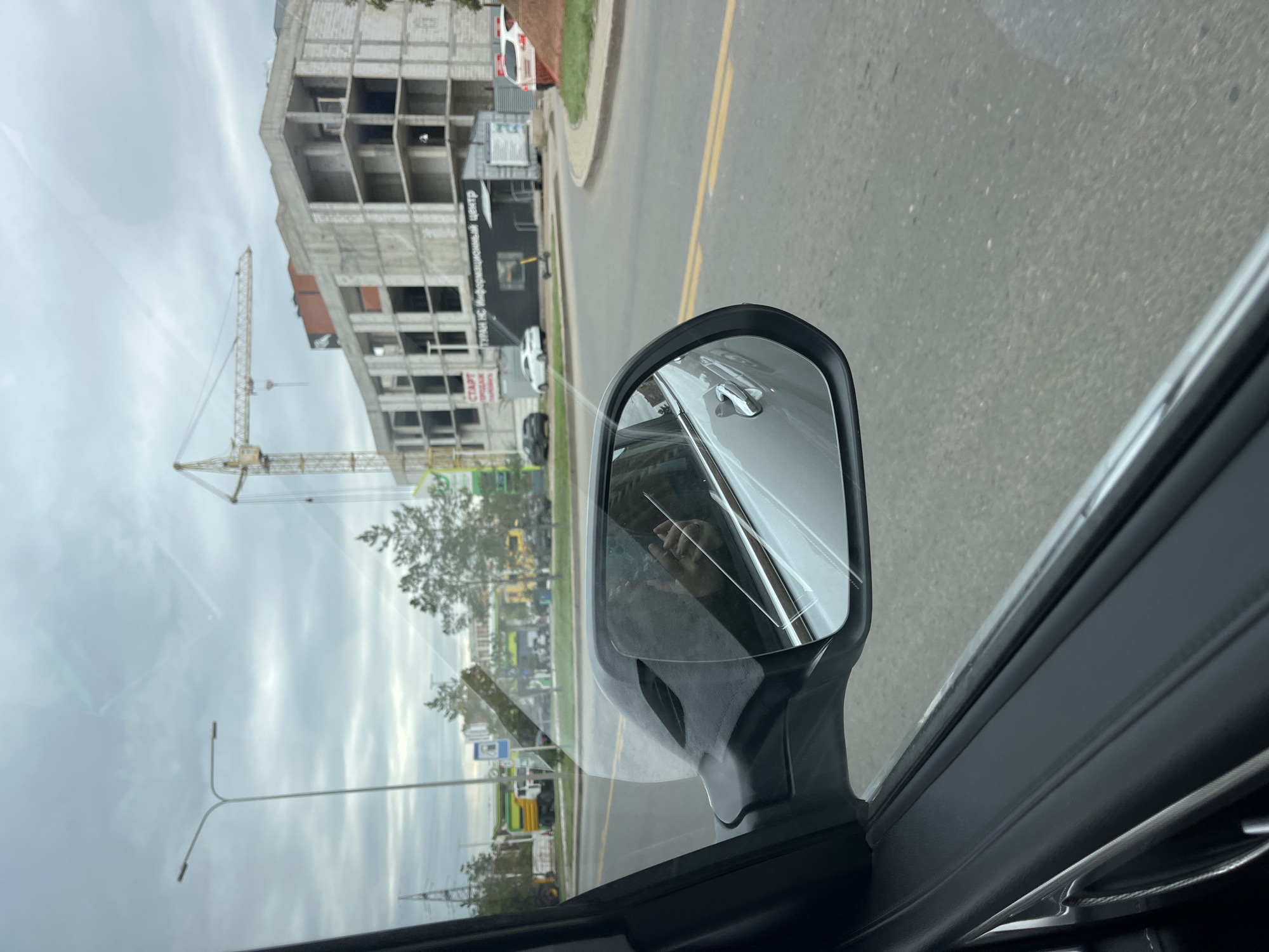 The image is taken from inside a vehicle, looking out the side window and reflecting in the side mirror. In the foreground, the interior of the car is visible along with the side mirror. The reflection in the side mirror shows a person holding a phone.
The exterior scene shows a street with a construction site. A large concrete building under construction is prominent, with a yellow crane positioned next to it. There are other vehicles on the road, including cars and a truck. A gas station with visible signage is also present in the background, along with trees and streetlights. The sky is overcast with clouds.
The text on the building reads "ТУРАН НС Информационный Центр", which translates to "TURAN NC Information Center". This indicates the location is likely in Astana, Kazakhstan, as "Turan" is a historical and geographical term often associated with Central Asia, and the construction of modern buildings is common in Astana. The street appears to be a multi-lane road with yellow lane markings. The overall environment suggests an urban setting undergoing development.