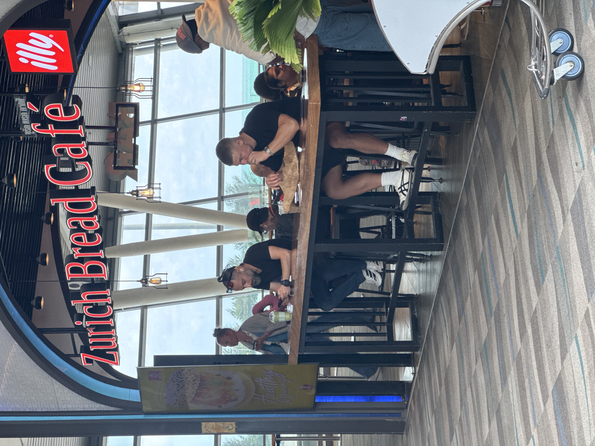 The image shows the interior of a Zurich Bread Café, located in Mai Khao, Thailand. The café features a long communal table where several patrons are seated. In the foreground, two men are sitting at the table. The man on the left is wearing a black t-shirt and jeans and is looking at his phone. The man on the right is wearing a black t-shirt and shorts and is eating something from a paper bag. Further down the table, other individuals are visible, including a woman in the background walking away from the table. The café is brightly lit, with large windows offering a view of the outdoors. The flooring is carpeted with a geometric pattern, and a luggage cart is visible in the lower right corner. The signage above the counter reads "Zurich Bread Café" in red lettering, with an "illy" logo to the right.