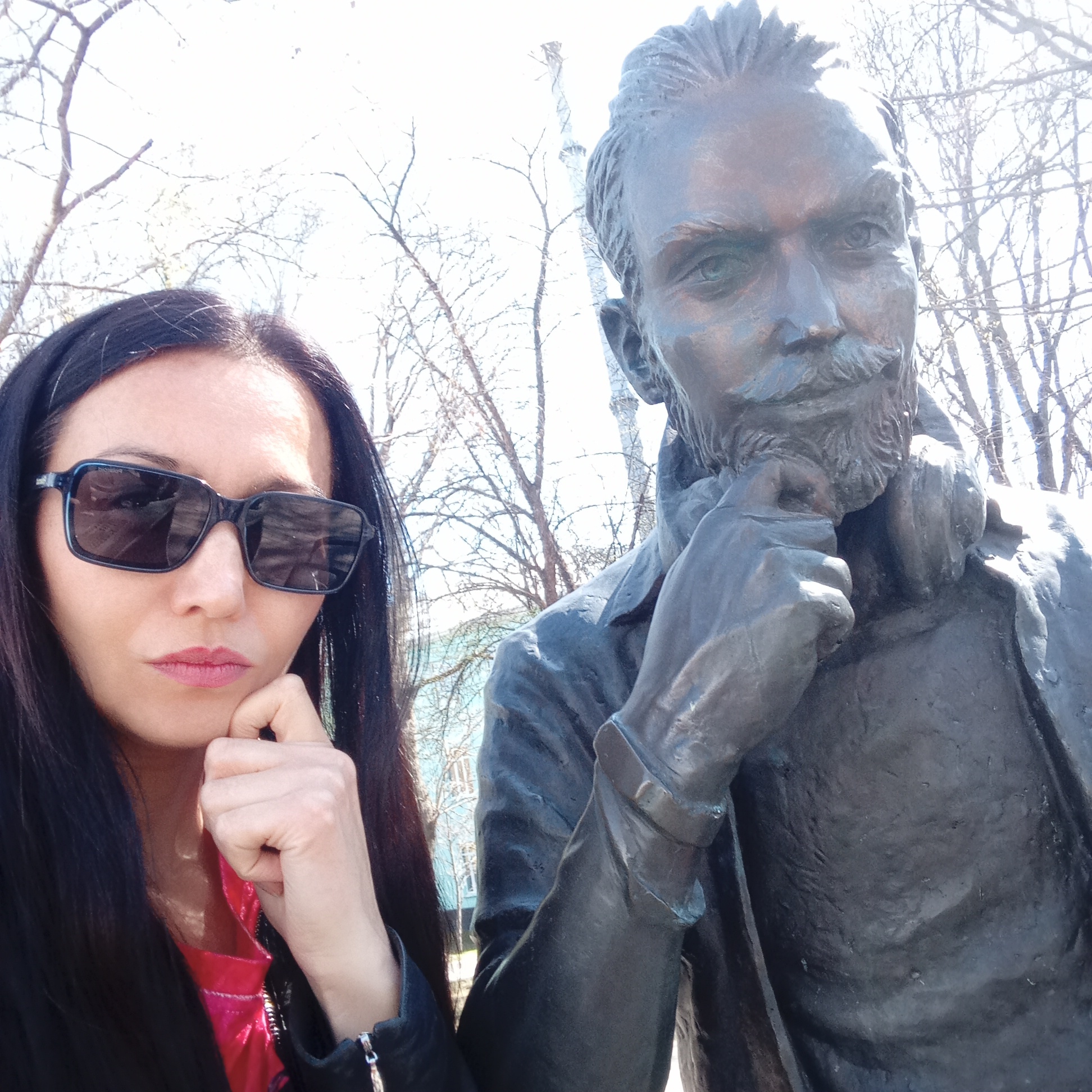 A woman in sunglasses and a black jacket poses beside a bronze statue of a man in Pervomaysky, Russia. Both the woman and the statue have their hands positioned as if they are in thought. The background consists of bare trees and a building.