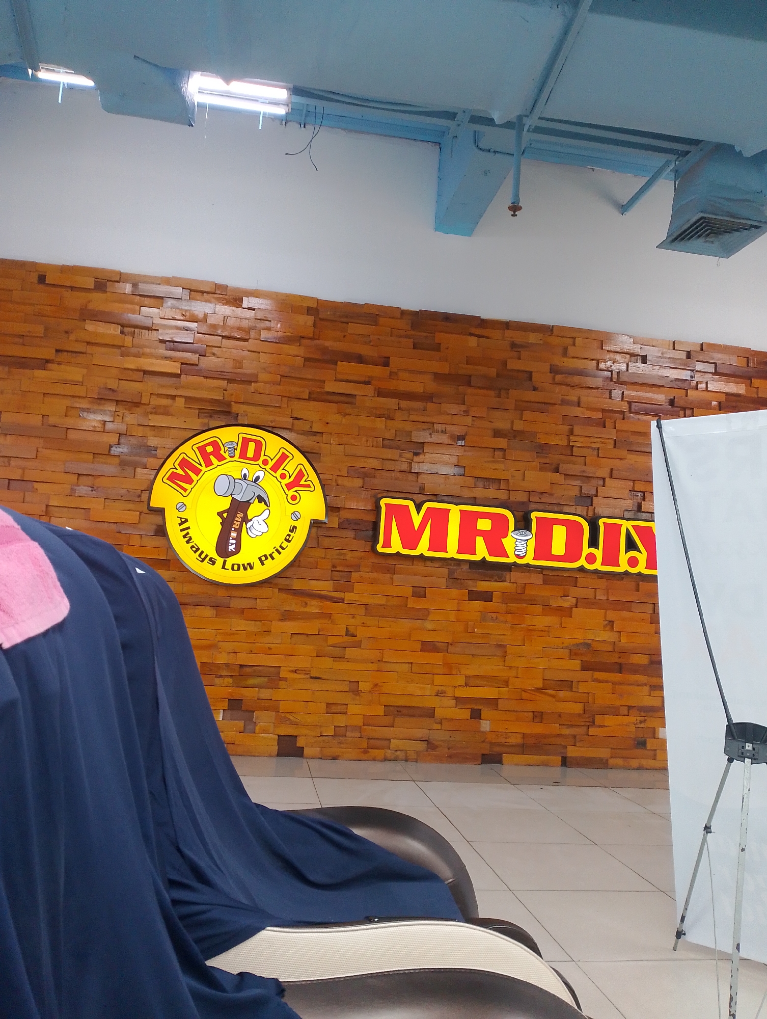 The image shows the interior of a MR. D.I.Y. store, a retail chain known for home improvement products, located in Batam, Indonesia. The store features a distinctive wooden plank wall as a backdrop for the MR. D.I.Y. signage. The signage is in two parts: one circular logo featuring a cartoon hammer and the text "MR. D.I.Y. Always Low Prices," and a rectangular sign with the text "MR. D.I.Y." The store's ceiling is painted blue and shows exposed ductwork and lighting fixtures. In the foreground, a portion of a large, upholstered chair covered with dark blue fabric and a pink towel is visible. To the right of the signage, a white advertising board on a tripod stand is present. The floor is tiled. The overall impression is that of a retail environment, likely a section dedicated to showcasing the brand.