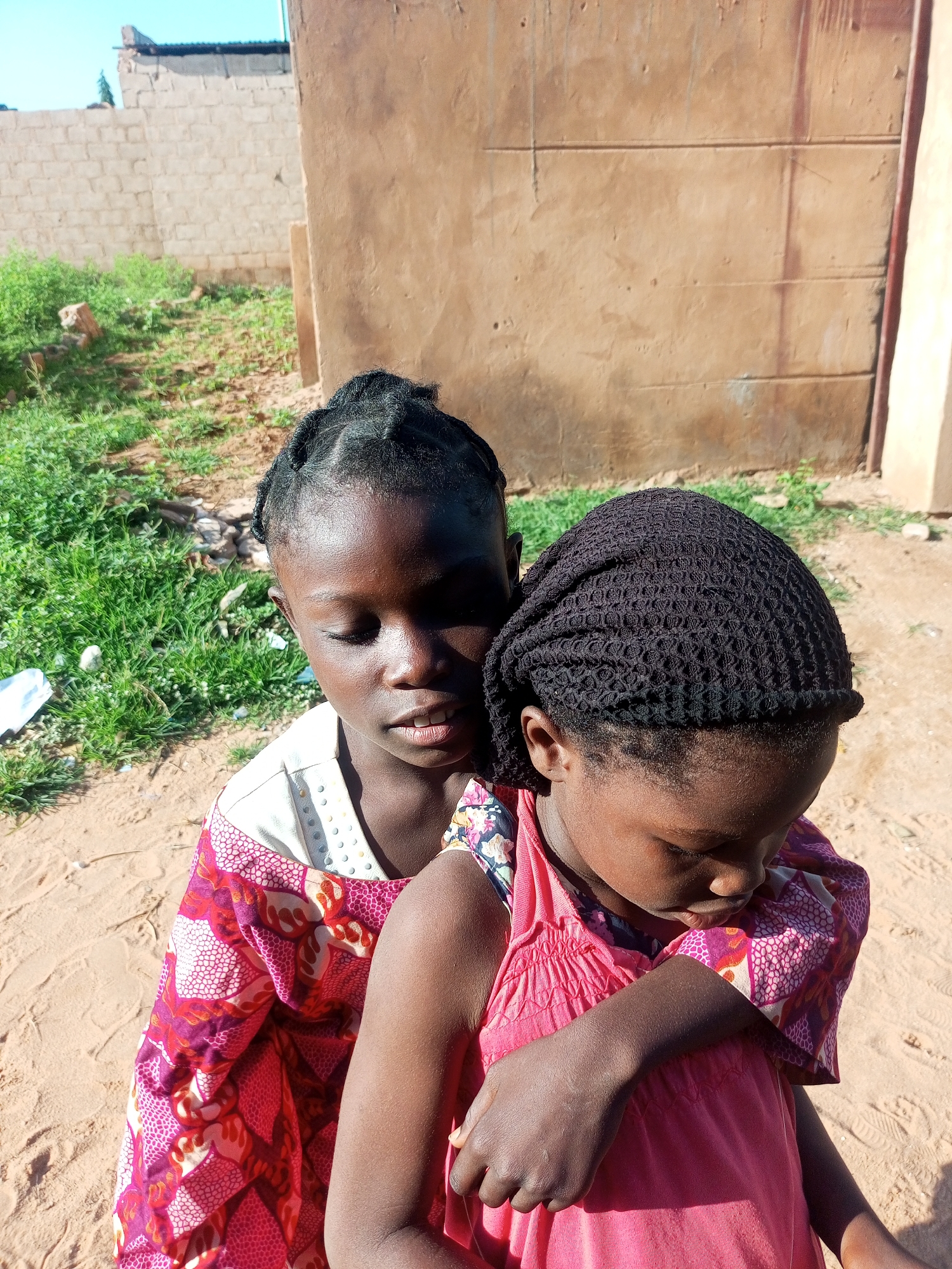 Two young Black girls stand close together in a sandy outdoor area, with a brown, stucco-like building and a low brick wall visible in the background. The girl in the foreground, on the right, wears a bright pink dress and has dark hair covered by a black, crocheted headscarf. Her head is bowed, and her arms are wrapped around the other girl. The second girl, on the left, is partly obscured. She has dark, braided hair and wears a patterned dress with pink, white, and red designs. Her head is also bowed, and she appears to be looking down. The ground is sandy and uneven, with patches of green grass and debris visible. The overall lighting suggests it is daytime, with sunlight casting shadows.