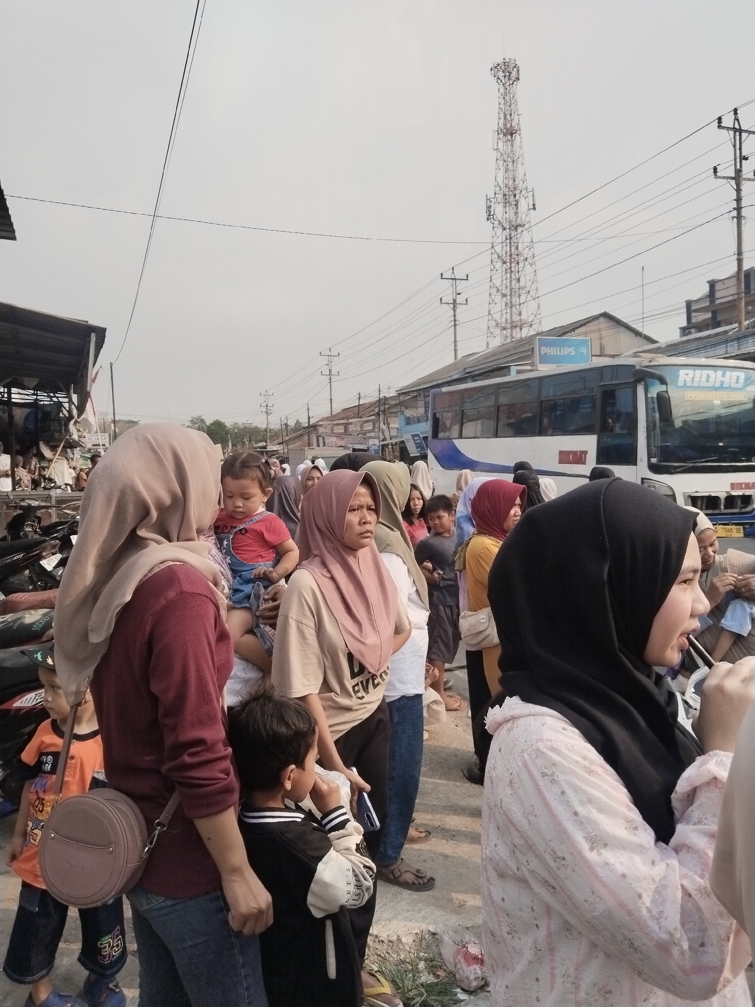 In Balapulang Kulon, Indonesia, a diverse crowd of people, including women in hijabs, men, and children, are gathered outdoors. They appear to be waiting or moving along a street, with some individuals holding or carrying others. In the background, a tall telecommunications tower is visible, along with a bus labeled "RIDHO" and buildings. The environment suggests a public area, possibly a market or transportation hub, with parked motorcycles also present. The sky is overcast.