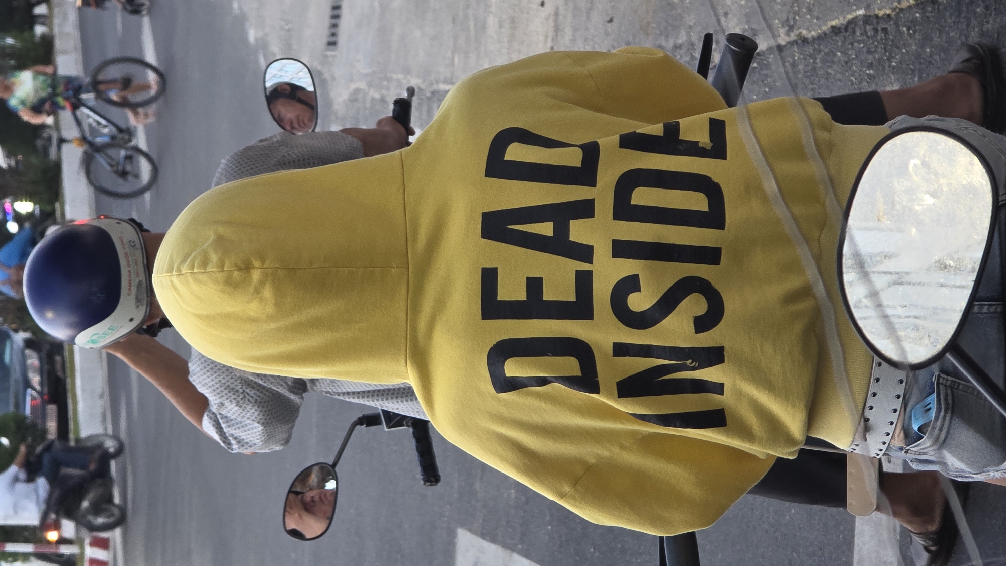 A person on a scooter in Bắc Nha Trang, Vietnam, wears a yellow hoodie with the words "DEAD INSIDE" printed in black letters on the back. Another person sits in front of them. Both are on a scooter, on what appears to be an avenue with other vehicles and pedestrians.