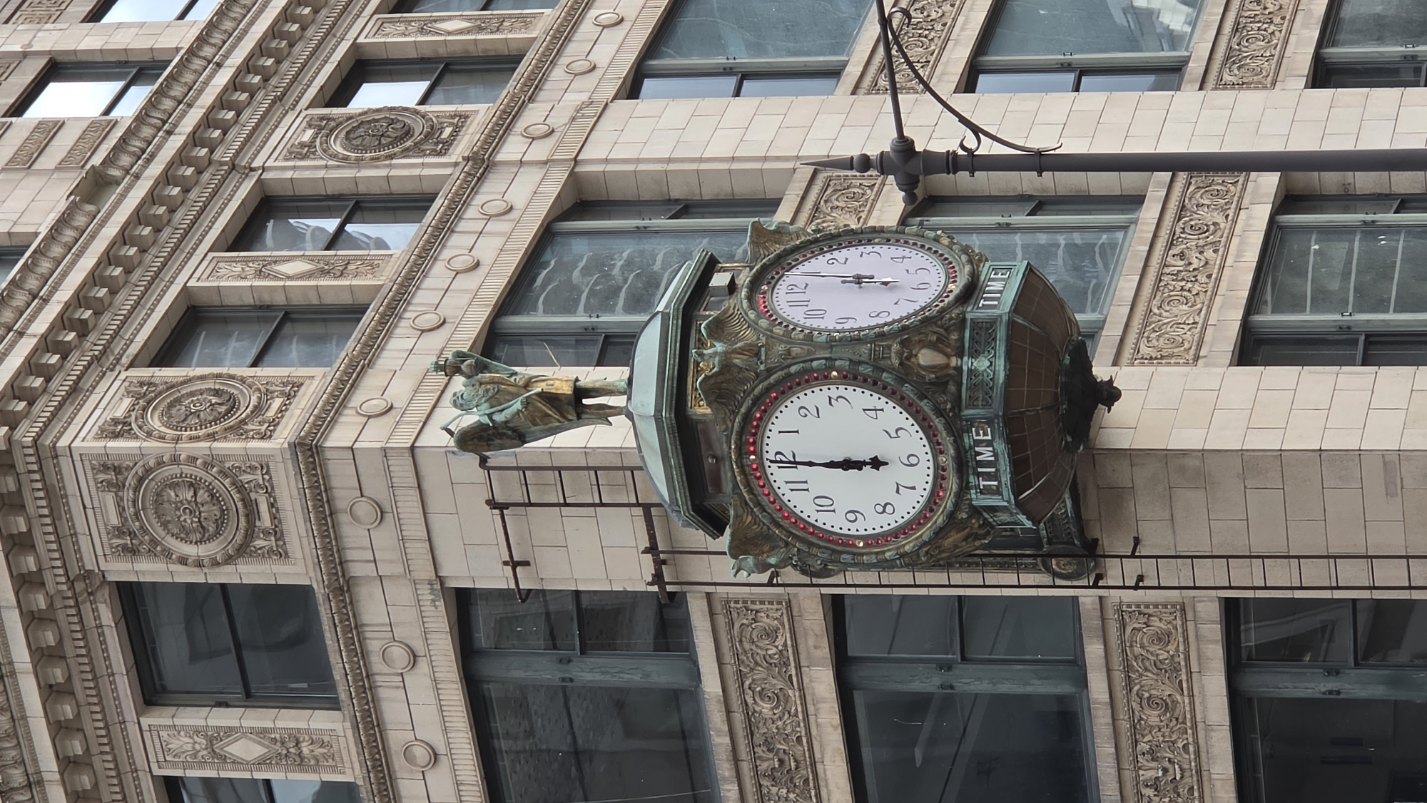 The image shows the northeast corner of the Wrigley Building in Chicago, Illinois, United States of America, focusing on the exterior architectural details and a prominent double-sided clock. The building's facade is constructed of light-colored terra cotta with ornate carvings and decorative elements, including circular medallions and relief patterns. Multiple windows, some with dark panes and others reflecting the sky, are visible on various levels.
Mounted on the corner of the building, suspended from a metal structure attached to the facade, is a large, ornate, double-sided clock. The clock faces are white with black Arabic numerals and hands. The left clock face shows the time as approximately 12:00, with both hands pointing to the 12. The right clock face is partially obscured, but appears to show a time close to 12:40. The clock is framed by decorative bronze elements, including winged figures and a crown-like top. A golden statue of an angel holding a book and a lamp stands atop the clock. Below the clock faces, the word "TIME" is inscribed in capital letters on each side. A dark metal ladder is attached vertically to the building wall next to the clock. A portion of a dark metal lamppost with decorative scrollwork is visible to the right of the clock. The overall scene depicts a historic architectural landmark in an urban environment.