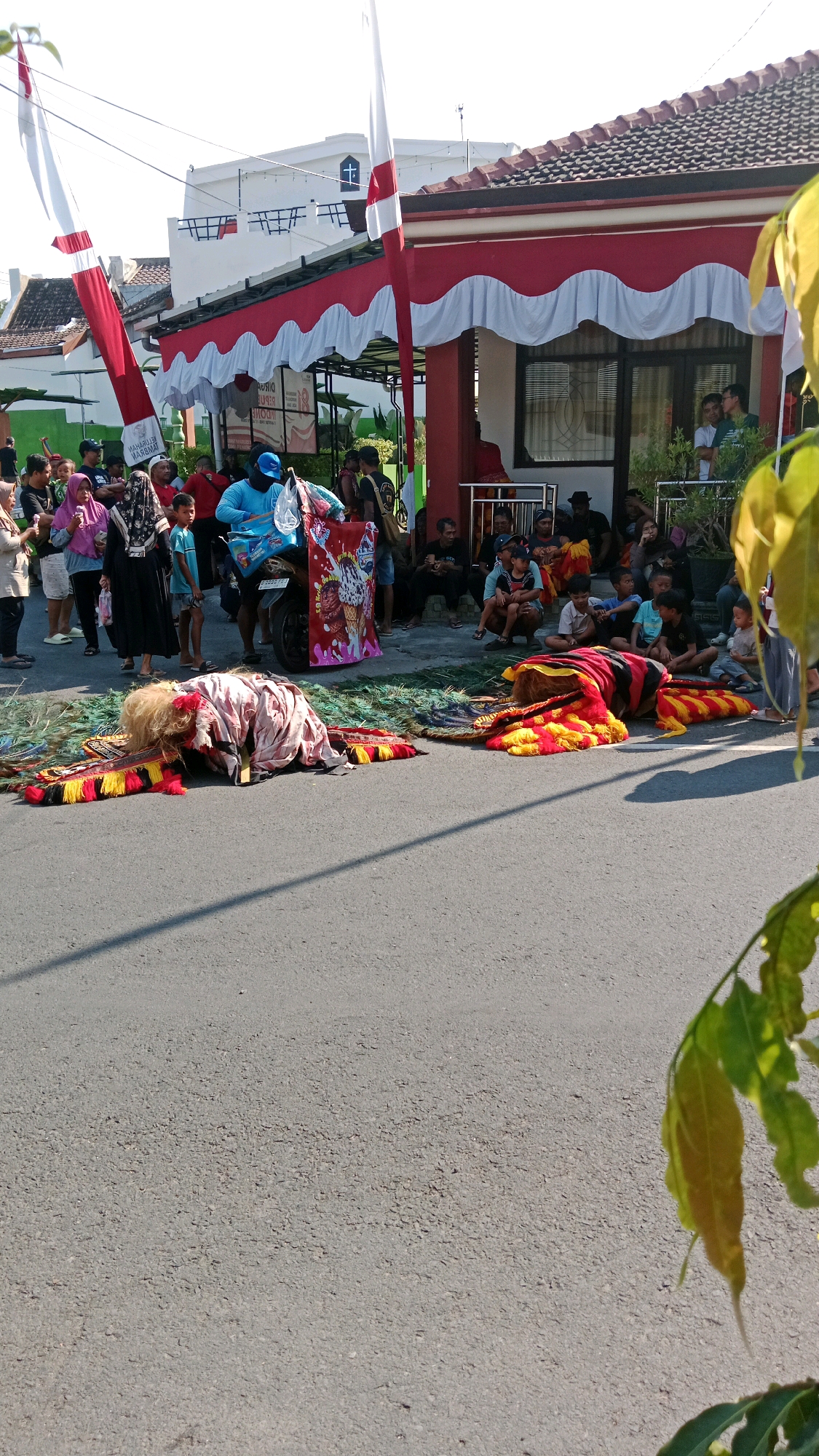 In Magetan, Indonesia, a street scene unfolds with a mix of cultural performance and community gathering. Two elaborate costumes, resembling mythical creatures with vibrant red, yellow, and black tassels and feathery accents, lie on the asphalt, one partially obscured by the other. Surrounding these, a teal-colored mat is laid out, possibly for the performance.
A crowd of onlookers, including men, women, and children, are gathered on the side of the road, some sitting on the ground, others standing. They are dressed in casual attire, suggesting a local event or festival. A motorcycle, adorned with a large, colorful banner featuring an ice cream cone illustration, is parked near the performers' costumes.
Behind the crowd, a building with red and white awnings and a tiled roof provides a backdrop. Red and white Indonesian flags are prominently displayed, fluttering in the breeze from tall poles, indicating a possible national holiday celebration. The environment appears to be a typical urban street, with a paved road and sidewalk, under a bright, sunny sky.