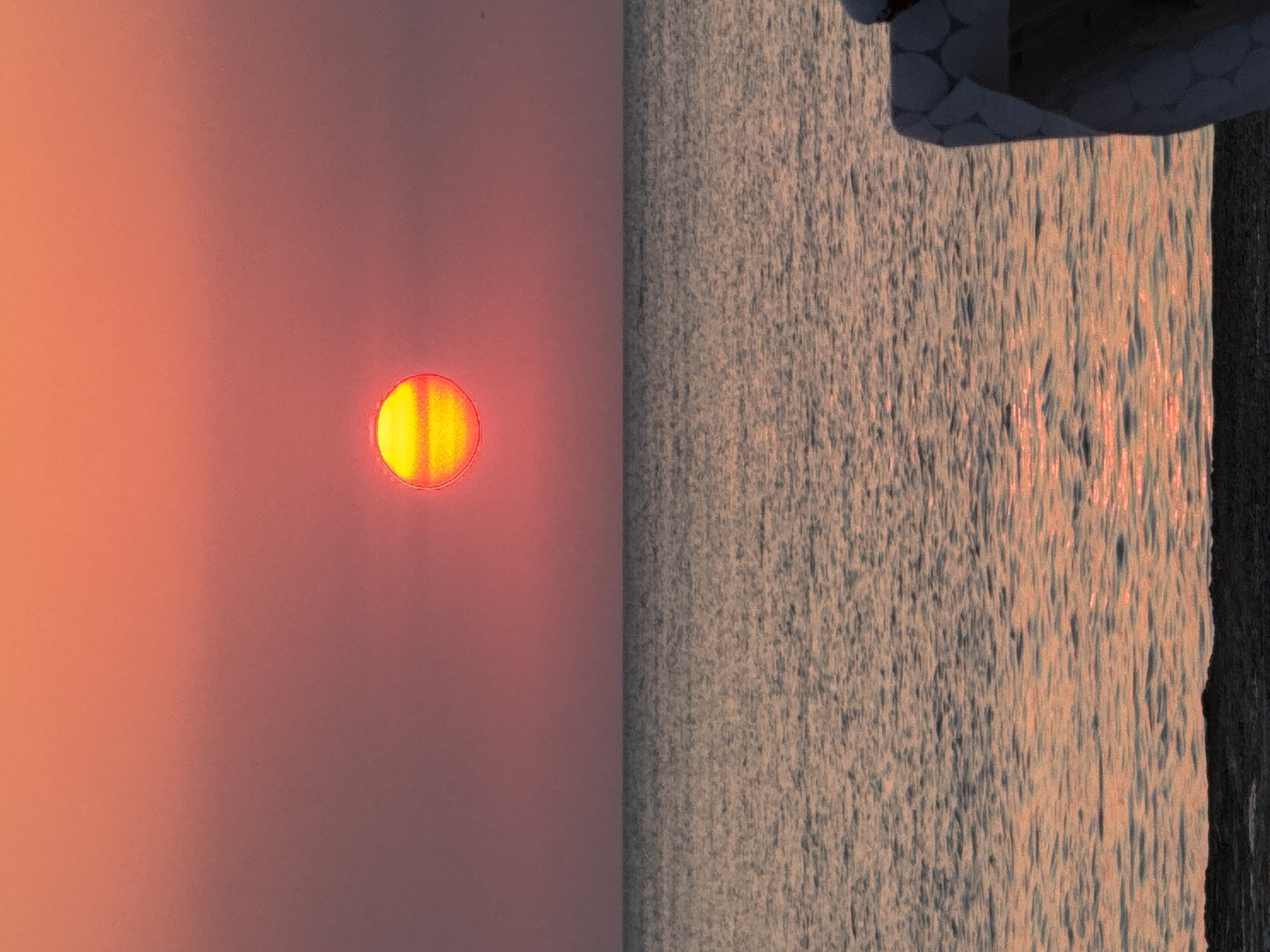 A close-up, vertical, eye-level shot captures the setting sun over a calm sea. The sun, a bright yellow disc with red horizontal bands, is positioned in the upper center of the frame, dominating the rosy-pink sky. The sea below reflects the warm hues of the sky, with gentle ripples creating a textured surface. In the foreground, the dark, pebbly shore is visible, and to the right, a portion of a white and grey patterned deck chair is seen from behind, partially obscuring the seascape. The image appears to be taken in Sevastopol, Ukraine.