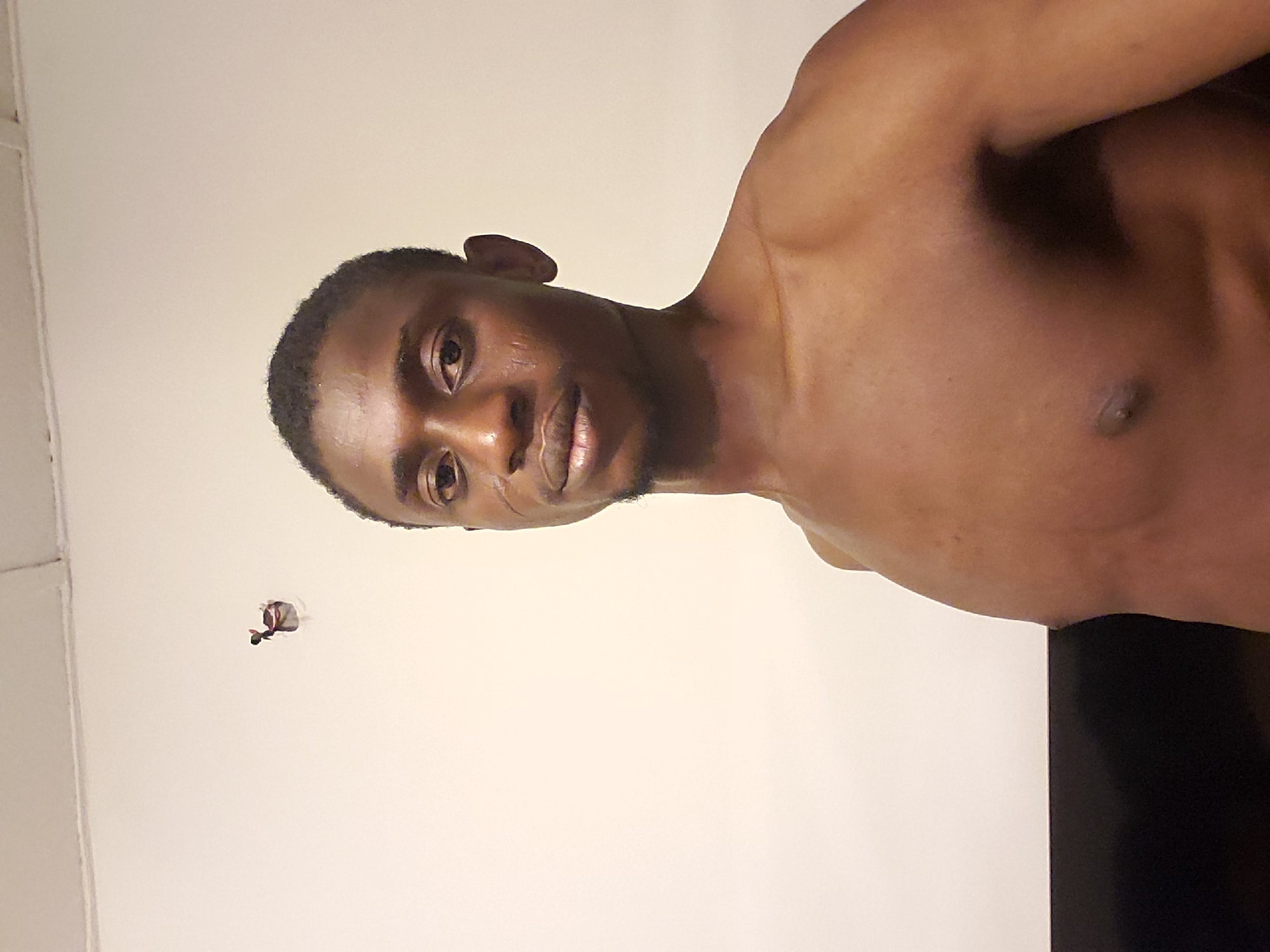 A medium shot depicts a shirtless, dark-skinned male posing indoors against a white wall. A wire protrudes from a hole in the wall. The setting's location in Adonkolo, Nigeria, is not evident from the image.
