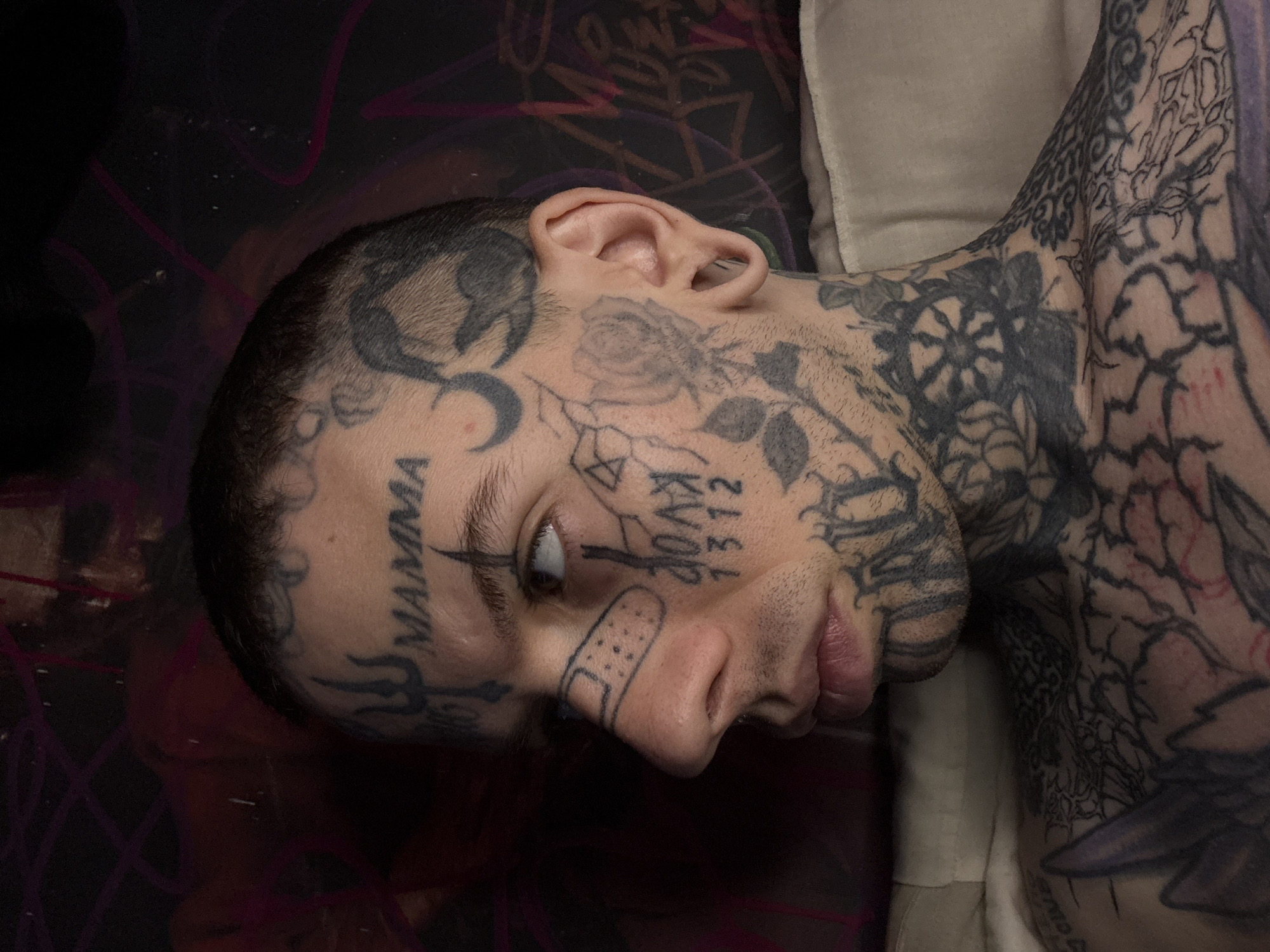 The image features a person with extensive black ink tattoos covering their face, head, neck, and upper body. The subject is seen from a slightly elevated side angle, lying down indoors, with their head turned to the right and eyes looking upwards. Their expression is neutral to contemplative, with lips slightly parted.
The face is densely covered in tattoos. On the left temple, the word "MAMMA" is tattooed vertically, and a crescent moon design is visible above the left eyebrow. A small, stylized band-aid tattoo is placed on the bridge of the nose. On the right cheek/jawline area, the word "KVAD" and the numbers "1312" are inked. Other designs, including floral patterns, geometric shapes, and abstract lines, cover the remaining facial skin, extending onto the very short or shaved scalp. The neck and visible portions of the chest and shoulders are also heavily adorned with intricate patterns, including what appears to be a spiderweb, roses, and various abstract designs.
The person has pale skin and visible stubble on the chin and cheeks. The background behind the head is a dark, patterned surface, possibly a blanket or pillow, with faint purple lines. A plain light-colored pillow or cushion supports the person's right shoulder. The lighting suggests an indoor environment. There are no direct visual cues within the image to indicate the location, but the provided context states it is Buenos Aires, Argentina.