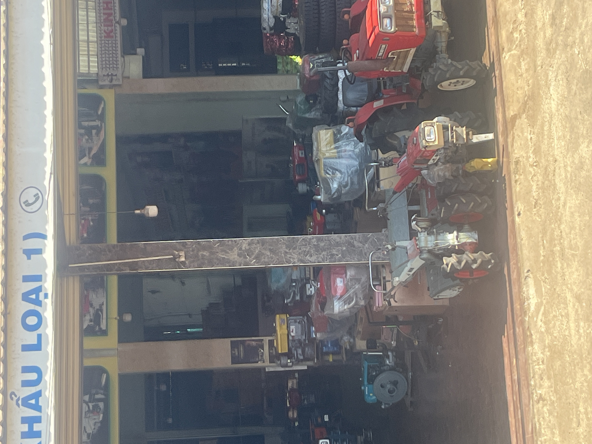 The image captures a storefront in Trung Hồ, Vietnam, which appears to be a dealer of agricultural machinery. A large sign above the entrance reads "KHÂU LOẠI 1)" in blue lettering. A telephone icon is also visible on the sign.
Inside the dimly lit shop, various agricultural machines are displayed. In the foreground, two red multi-purpose tractors are prominently featured. The tractor on the right has "YN 20100" written on its front. To the left of these tractors, a turquoise water pump is visible. Further back, other machinery, including red engines and tires, are stored. The environment suggests a commercial setting for machinery sales.
