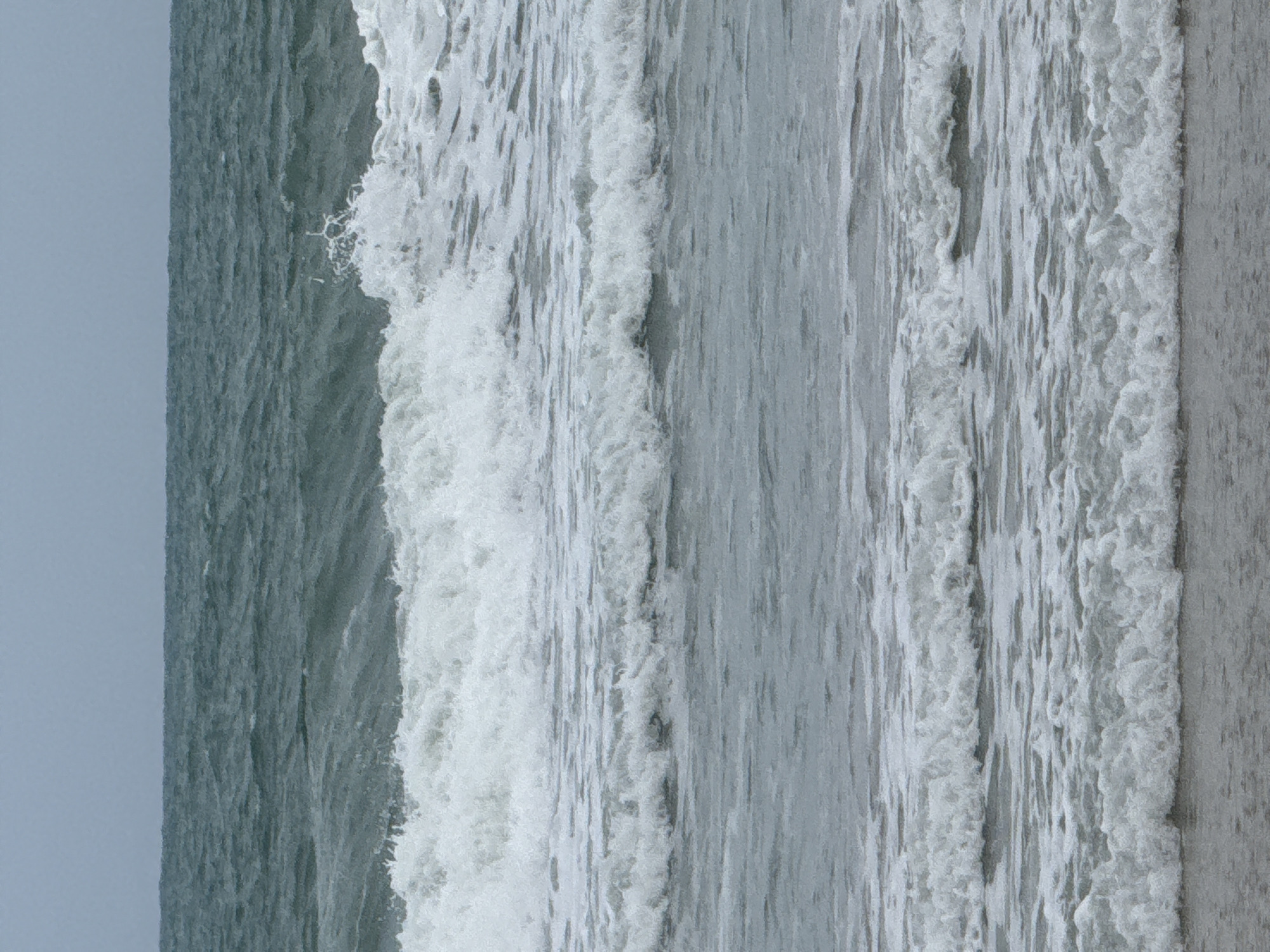 The image shows a view of the ocean from a beach. The sky is overcast and the water is choppy with several waves breaking. The waves are rolling towards the shore and the foam from the breaking waves is visible. The beach itself is not visible in detail, but the edge of the water can be seen at the bottom of the image. The image appears to be taken in a natural setting with no buildings or people visible. The location is Karon Beach, Karon, Mueang Phuket District, Phuket, Thailand.