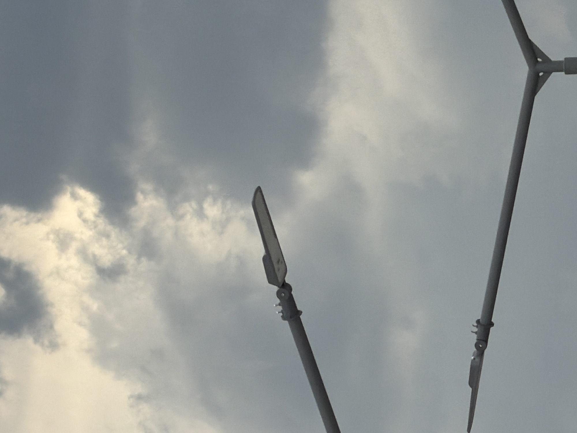 Two lampposts are visible against a cloudy sky. The upper lamppost has a gray pole extending diagonally from the left to the right, with a light fixture attached at the right end. The lower lamppost is a Y-shaped structure, also gray, with two arms extending horizontally. The sky is filled with gray and white clouds, suggesting overcast conditions. The image appears to be taken outdoors, likely in a public area where street lighting is present. The specific location in Căușeni, Moldova, is not discernible from the visual information alone. There are no people or discernible actions in the image.