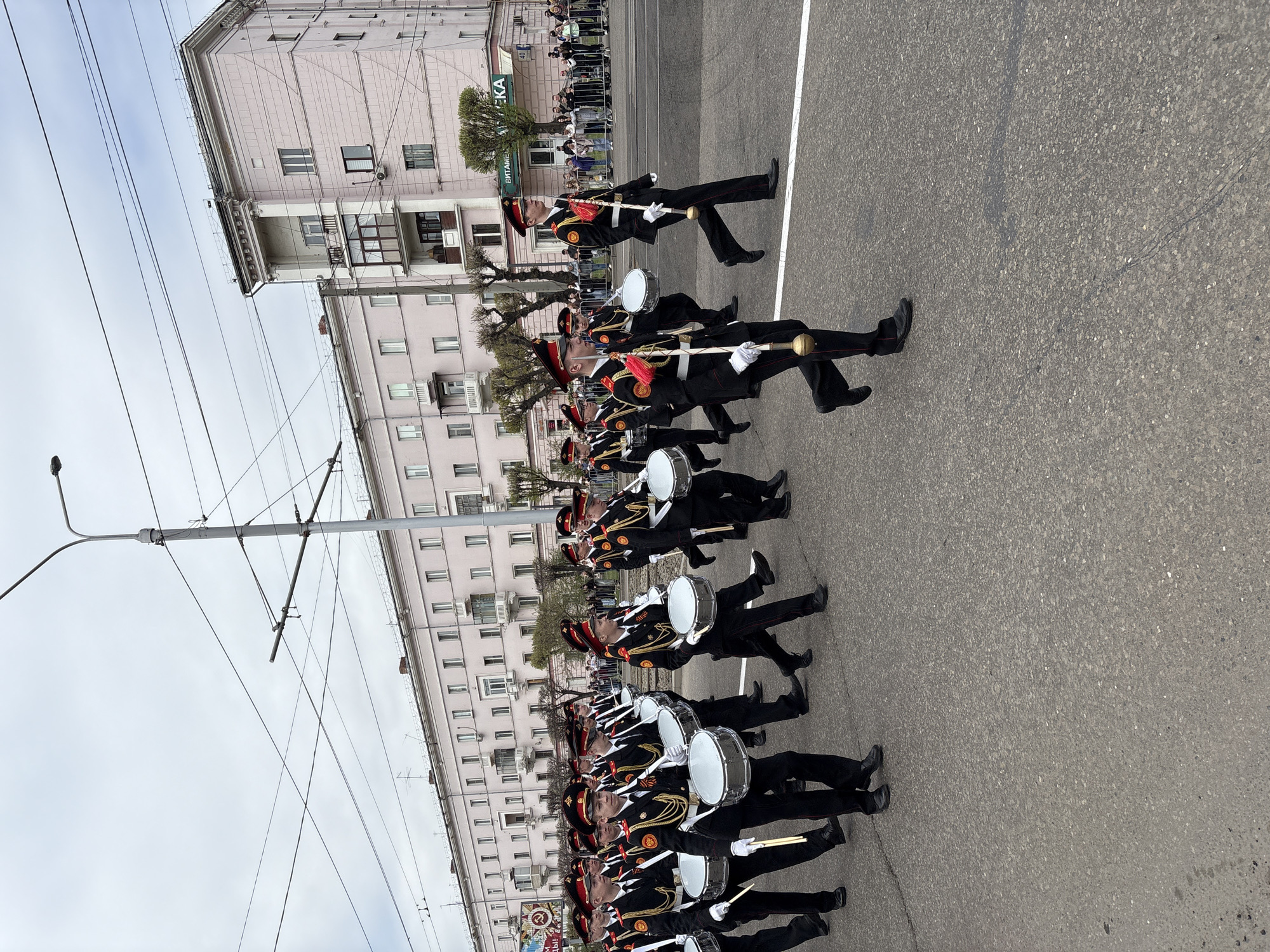 A military marching band in black uniforms and red hats is parading down a gray asphalt road in Unknown, Unknown. A large, multi-story building with light pink walls and white trim lines the left side of the road. Overhead power lines and a street light run parallel to the building. On the right side of the road, a crowd of people stands behind a white line, observing the parade. The sky above is overcast and gray.