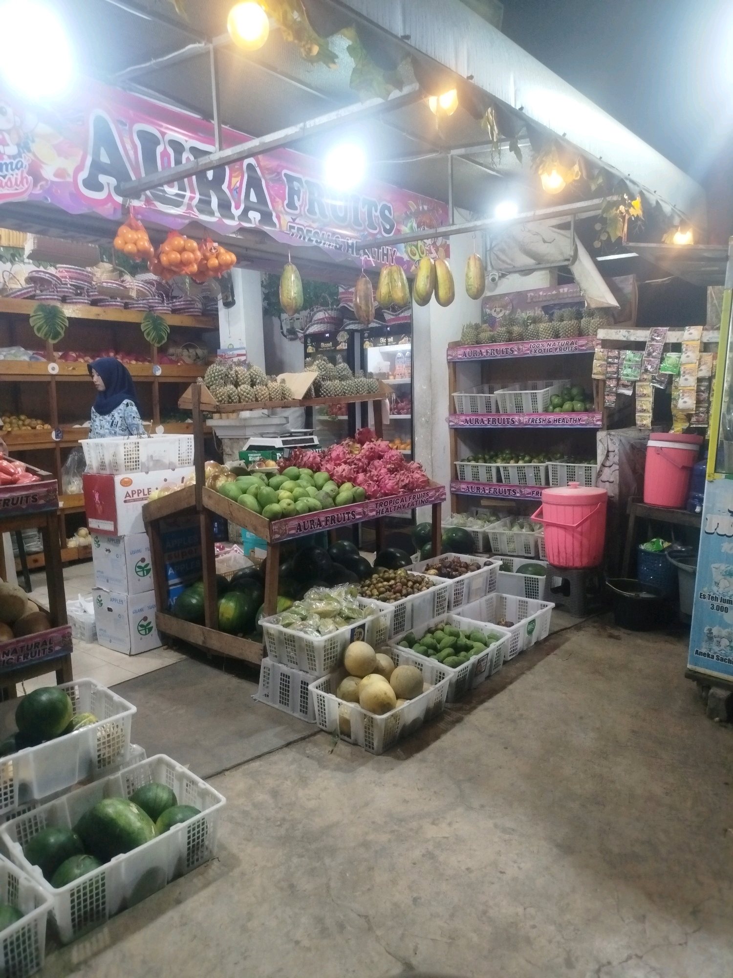 The image depicts a fruit stall, likely a market or a small shop, at night. The stall is brightly lit by overhead lights, illuminating a wide assortment of fruits displayed in white plastic crates and on shelves. In the foreground, several white crates are filled with green watermelons and other green produce. Further back, more crates contain a variety of fruits, including what appear to be avocados, melons, and possibly pineapples.
Behind the fruit displays, there are shelves stocked with more fruits, including large piles of dragon fruit and what looks like pineapples. A woman wearing a blue hijab and a patterned blue and white shirt stands behind a counter, partially obscured by boxes. The boxes are labeled "APPLE" and "GUANG YUAN."
Above the stall, a large banner with the text "AURA FRUITS" is visible, along with some hanging oranges and papayas. The environment appears to be an outdoor or semi-outdoor market setting, with a concrete floor and a simple roof structure. The overall impression is of a busy, well-stocked fruit vendor operating in the evening.
Based on the context of the prompt, this scene is located in the city of Burneh, Indonesia.