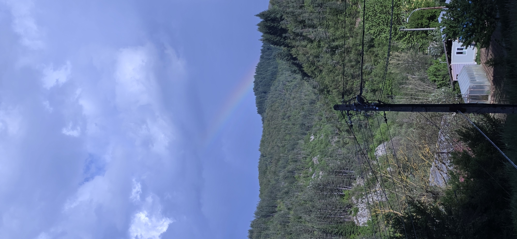 The image shows a rainbow in the sky, ending at the edge of a forested hillside. Utility poles with wires are in the foreground. In the background, a building and greenhouse are visible. The scene is located near Rudozem, Bulgaria.
