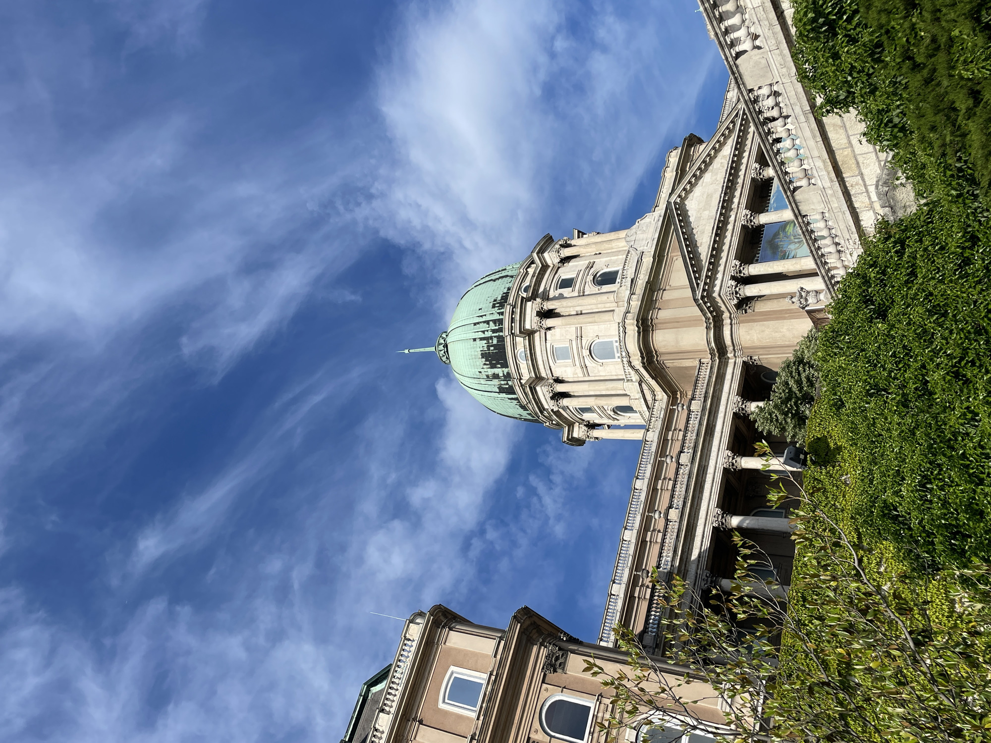 This is a low-angle shot of the Buda Castle Royal Palace in Budapest, Hungary. The image emphasizes the palace's dome, which is green, against a blue sky with wispy clouds. The building's architecture is visible, showing classical elements, including columns and decorative facades. Green foliage, likely ivy, partially covers parts of the building, adding to the overall aesthetic.