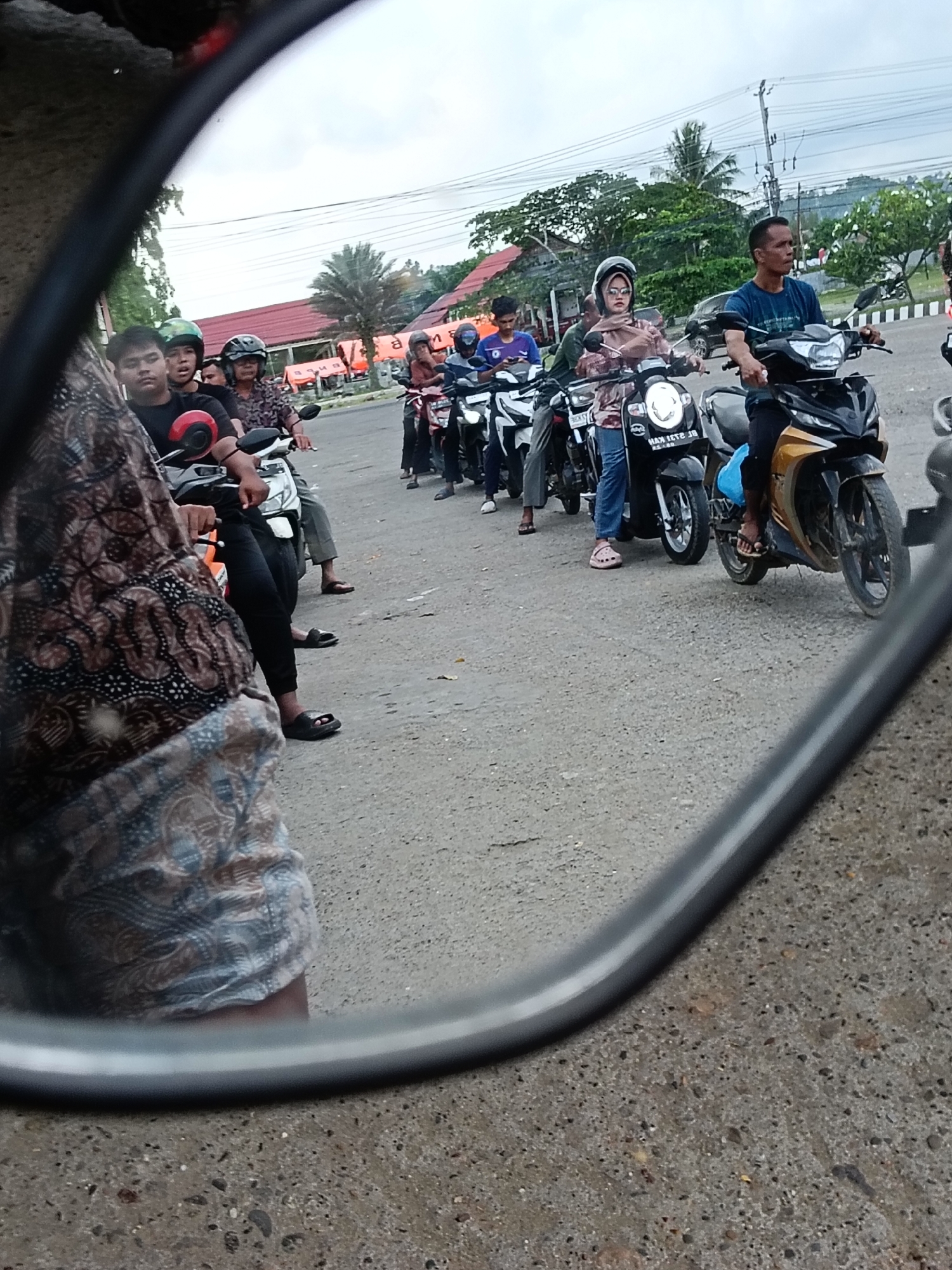 The image, likely taken from a vehicle's side mirror, shows a line of people on motorbikes waiting on a paved road. The location is possibly in Lhokseumawe, Indonesia. The people are wearing casual clothing, some with helmets. The motorbikes are a mix of different styles and colors. A building and trees are visible in the background. The overall lighting suggests it is daytime, but possibly overcast.