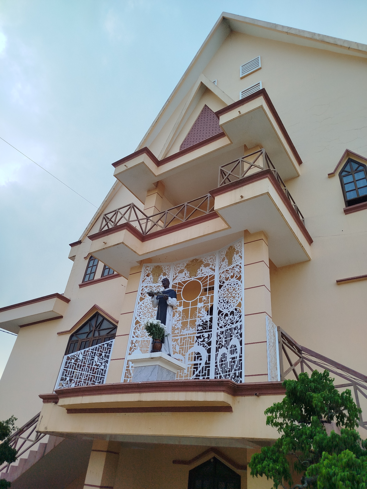 The image depicts the facade of a building, likely a church, located in Lạc Nghiệp, Vietnam. The building is multi-tiered with a light yellow exterior. A statue of a man in dark robes stands on a balcony, framed by a white, ornate screen. The building has multiple balconies with brown railings. Windows are visible on the side. A green tree is located near the base of the building. The sky is overcast.