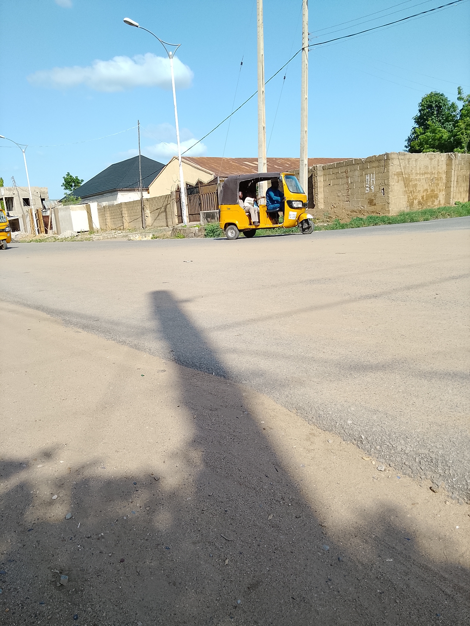 A yellow auto-rickshaw with two occupants is traveling on a paved road in Wauro Leggal, Nigeria. The auto-rickshaw is positioned in the right-center of the frame, moving from left to right. The driver, a man wearing a blue shirt, is at the helm. A passenger, also a man, is seated next to him. The auto-rickshaw has a dark canopy. The road surface appears to be asphalt. To the right of the road, a low, rough brick wall forms a boundary. Behind the wall, the landscape features scattered buildings, including one with a dark, sloped roof, and some vegetation. Power lines are visible suspended in the clear blue sky, supported by tall poles. The sun is casting a long shadow of the auto-rickshaw and a nearby person onto the road in the foreground. The overall environment suggests an urban or semi-urban setting with clear weather conditions.