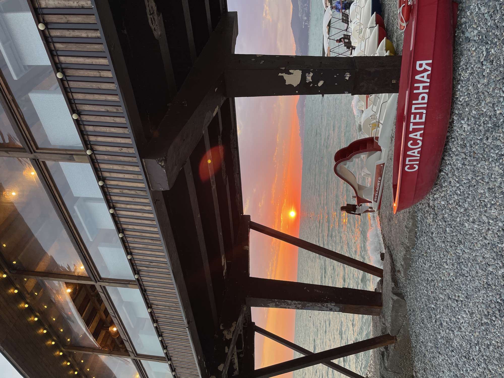 The image depicts a picturesque sunset over the sea, viewed from under a wooden pier. The pier's structure, supported by large pillars, extends over a pebble beach. Several pedal boats are lined up on the beach, with a prominent red boat in the foreground bearing the Cyrillic inscription "СПАСАТЕЛЬНАЯ," meaning "Rescue." A lone figure stands near the water's edge, facing the setting sun. The sea is calm with gentle waves, and the sky is painted with vibrant hues of orange and pink. In the distance, a coastline with hills and buildings is visible. The setting appears to be a coastal town, likely Khostinskiy, Russia, given the context.