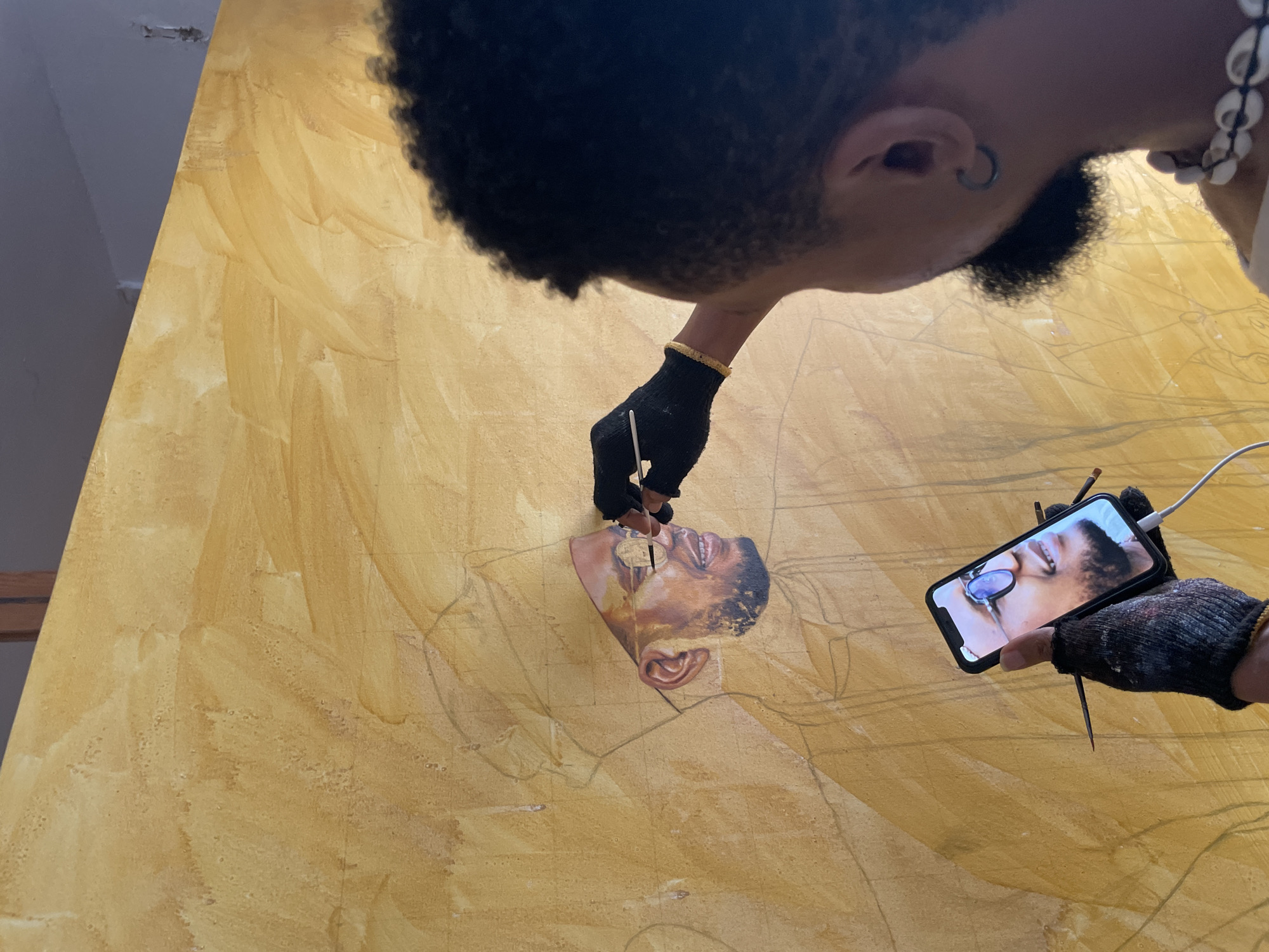 A low-angle, close-up shot captures a person painting a portrait. The individual, with short black hair and wearing a small hoop earring, is positioned above the canvas, head tilted downwards. The subject wears a beaded necklace and a gold bracelet. Gloved hands hold a thin brush, meticulously adding detail to the painted face on the canvas. The partial portrait is in warm brown tones. A smartphone displays a similar image of the person's face, presumably serving as a reference. The canvas is a light yellowish-brown with sketched outlines of the subject's body. The location is not discernible from the image, and there is no identifying information to specify the image location as Ado, Nigeria.