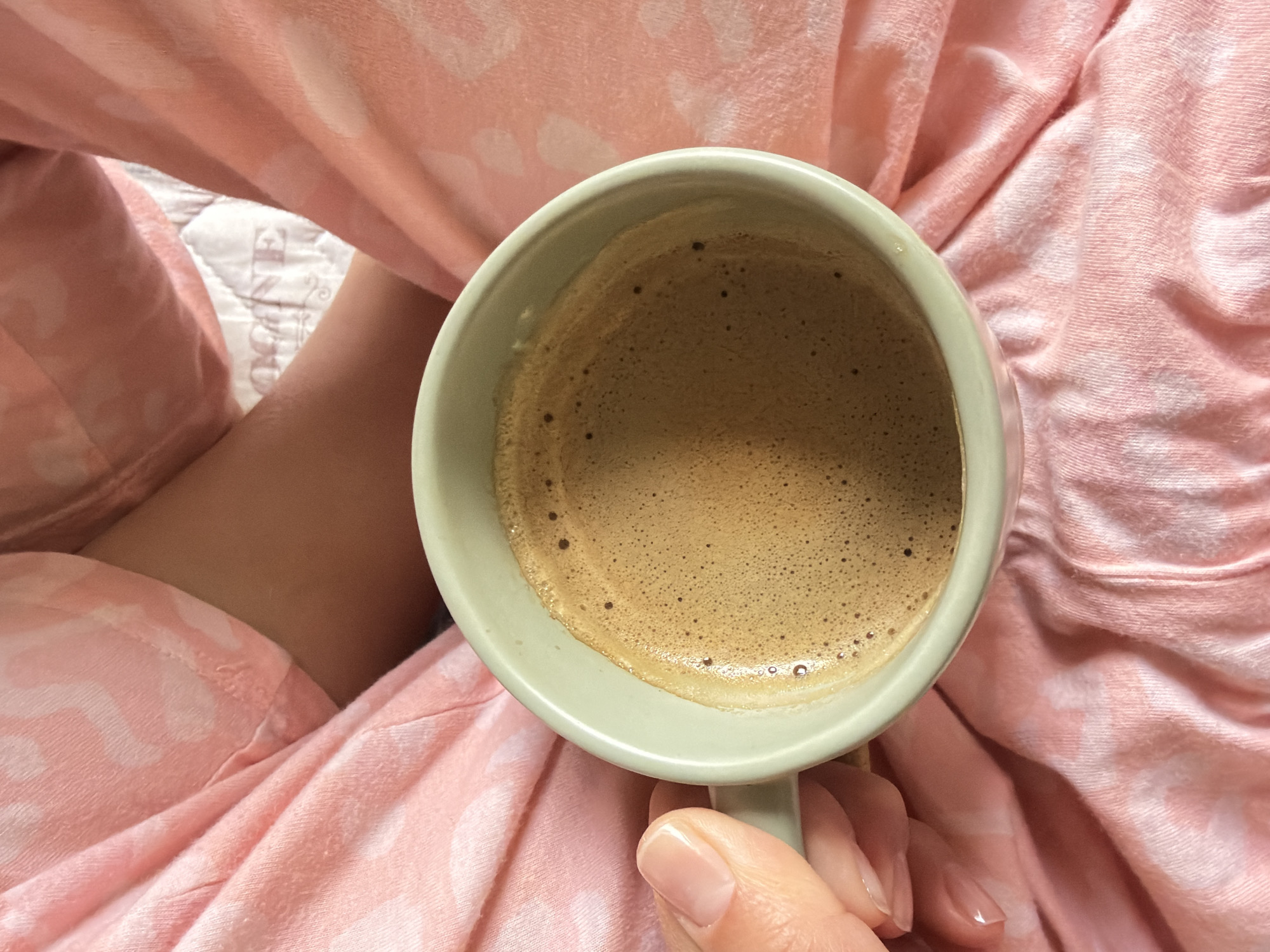 A close-up, eye-level shot shows a person holding a light green ceramic mug filled with brown coffee with foam. The person is wearing pink pajamas with white hearts. Part of their leg is visible under the pajamas. A small portion of a white quilted surface with a barely visible pattern is seen between the leg and the pajama pants. The photo provides no information regarding Pleven, Bulgaria.