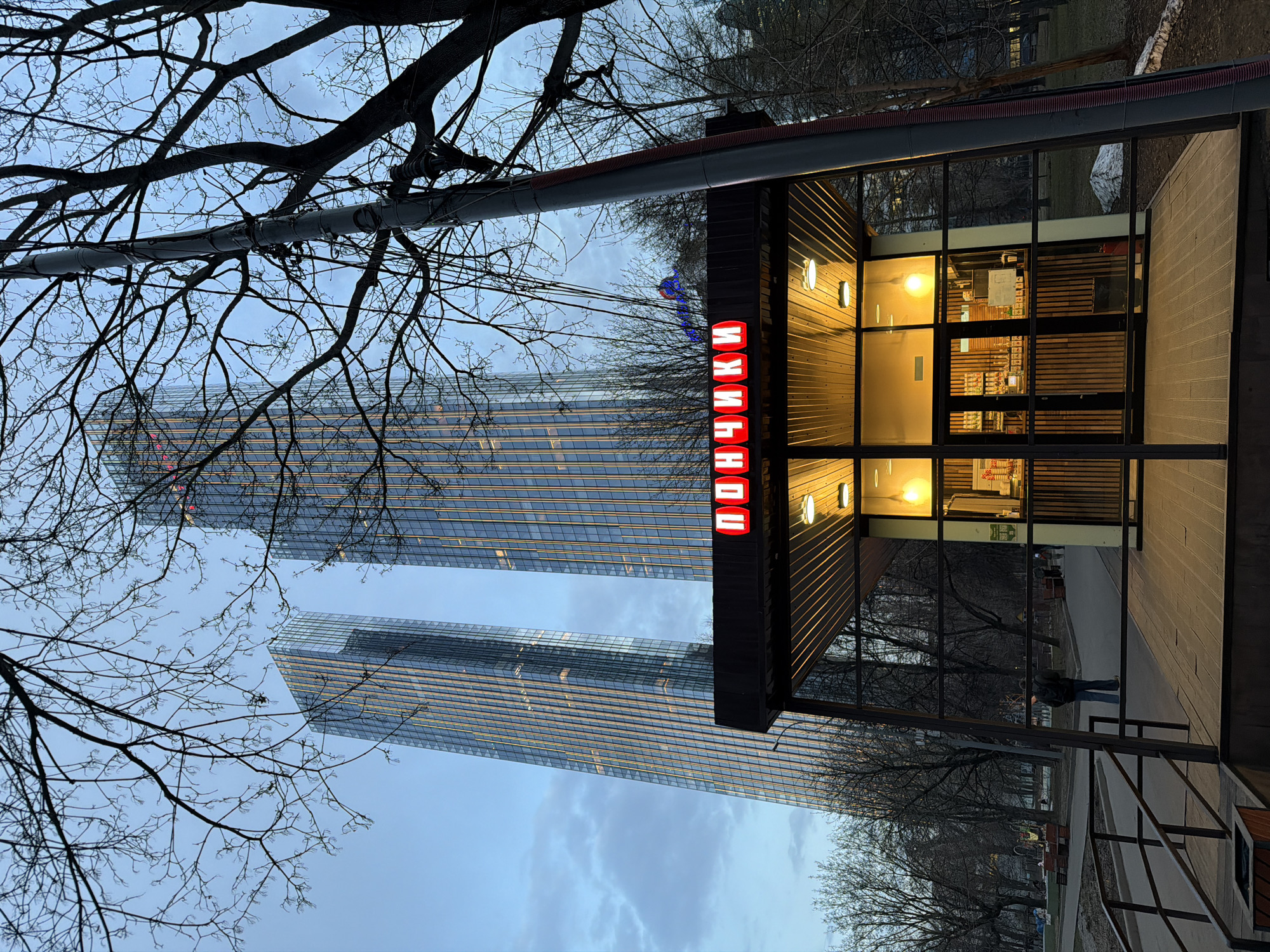 The image shows a "Donut" shop in Moscow, Russia, with skyscrapers in the background. The shop has a brown frame, a red illuminated sign with the word "Donuts" in Russian, and glass walls, with interior lighting turned on. The street outside appears empty, with a person walking in the distance on the right. In the foreground are bare tree branches. The sky is overcast. A support pole is in the scene.