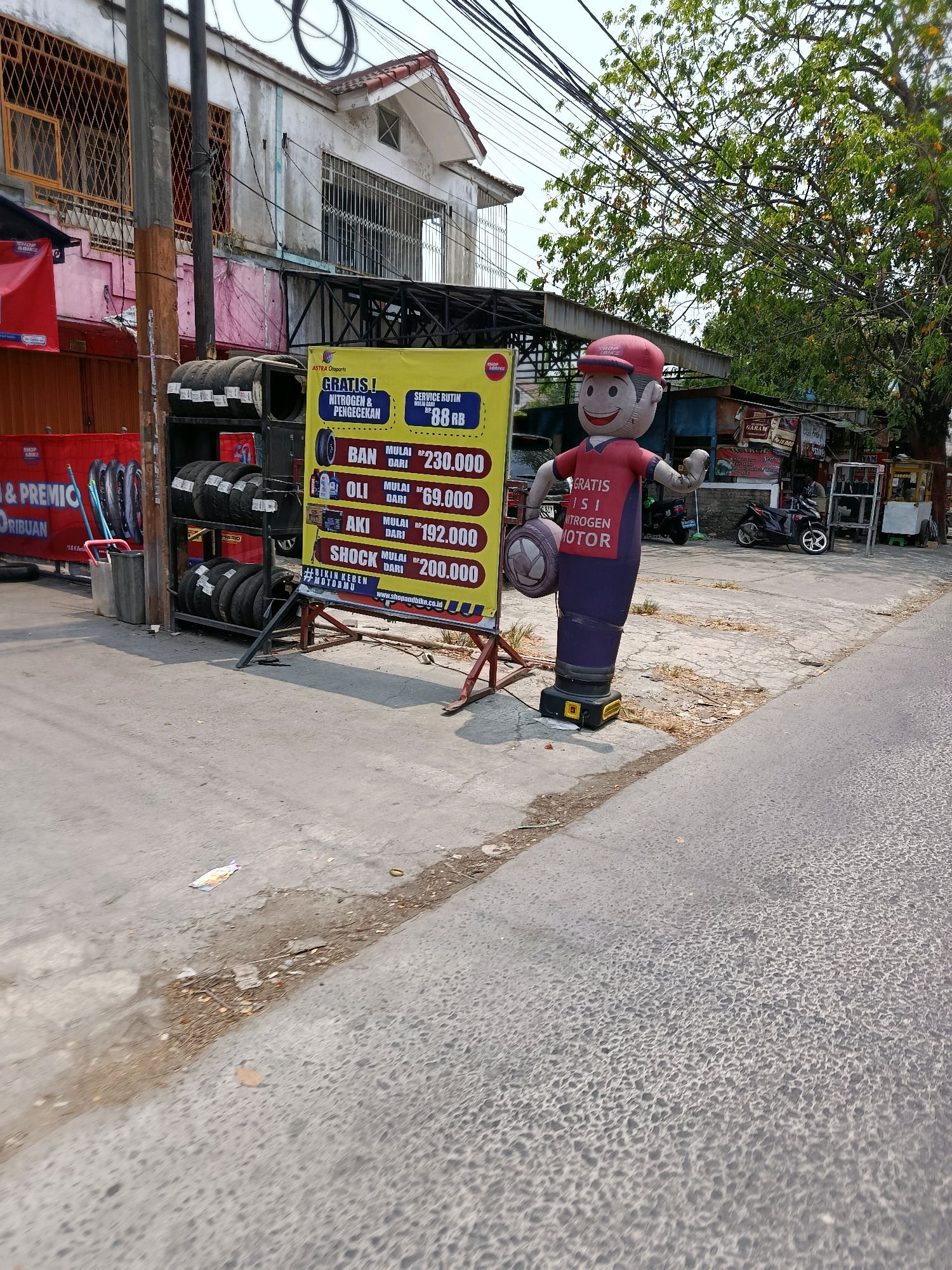 In Bekasi, Indonesia, a street scene unfolds with a prominent inflatable advertising figure standing on the sidewalk. The figure, dressed in a red shirt and purple pants, holds a tire and promotes "GRATIS ISI NITROGEN MOTOR" (Free Nitrogen Filling for Motorcycles). To its left, a yellow banner displays prices for motorcycle parts and services, including "BAN" (Tires) starting from Rp. 230,000, "OLI" (Oil) starting from Rp. 69,000, "AKI" (Battery) starting from Rp. 192,000, and "SHOCK" (Shock Absorbers) starting from Rp. 200,000. The banner also mentions "SERVICE RUTIN" (Routine Service) for Rp. 88,000. A rack of tires is displayed next to the banner. In the background, a multi-story building with barred windows and a tiled roof is visible, alongside other storefronts and parked motorcycles. The street is paved with asphalt and shows signs of wear and tear, with some debris on the side. Trees with green foliage are present in the background, and the sky is overcast.