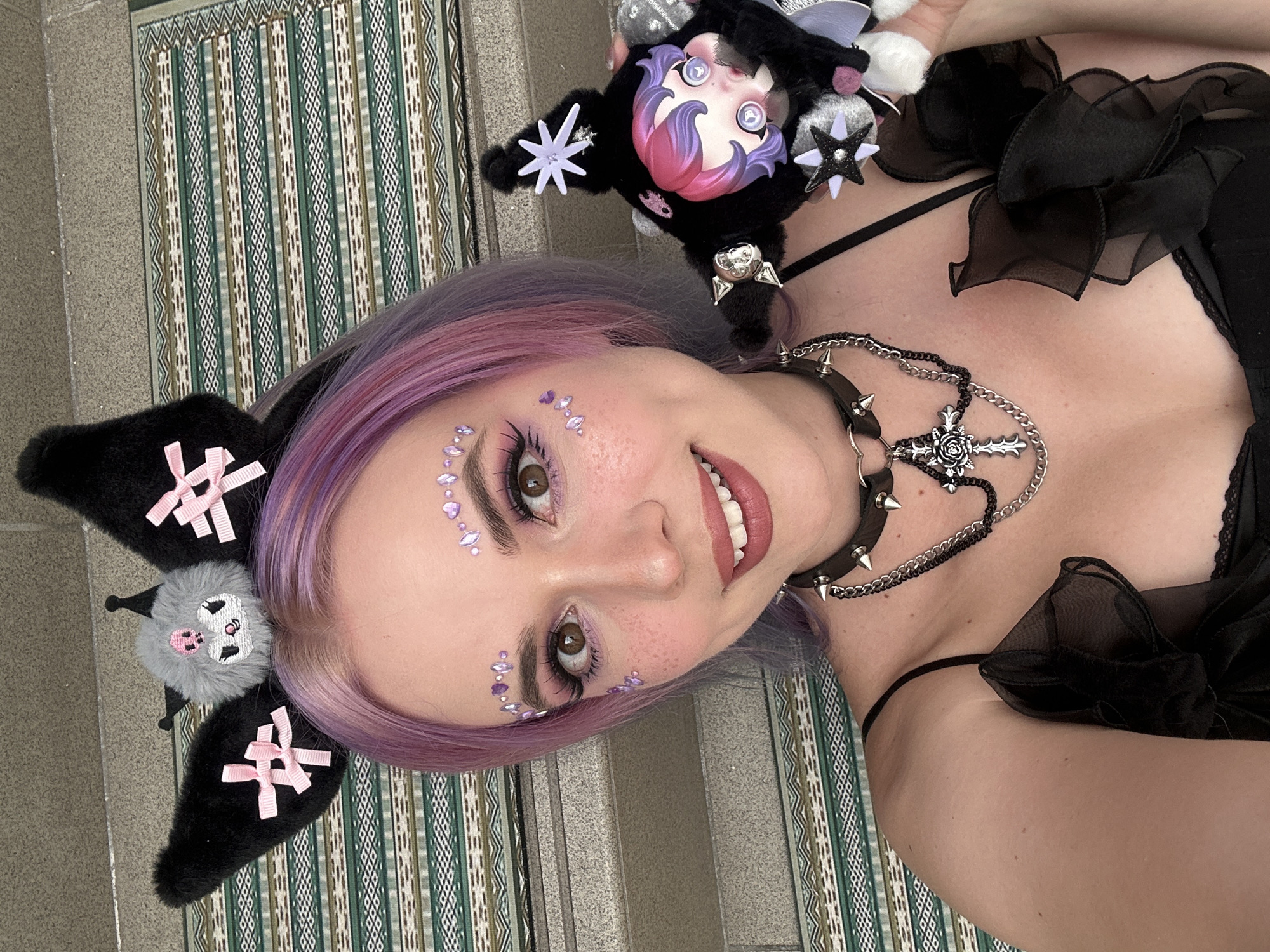 A young woman with lavender hair is posing for a selfie. She is wearing a black choker with spikes and a cross pendant necklace. There are small purple gemstones applied around her eyes as part of her makeup. On her head, she is wearing a headband with black plush ears adorned with bows and a small gray creature in between. She is holding a black plush doll with a stylized anime-style face, complete with stars attached to it. She is wearing a black top with ruffled accents. The location seems to be inside a building, as indicated by the patterned flooring and the wall in the background. The location details of City of Zagreb, Croatia are not present and cannot be extracted from the image.