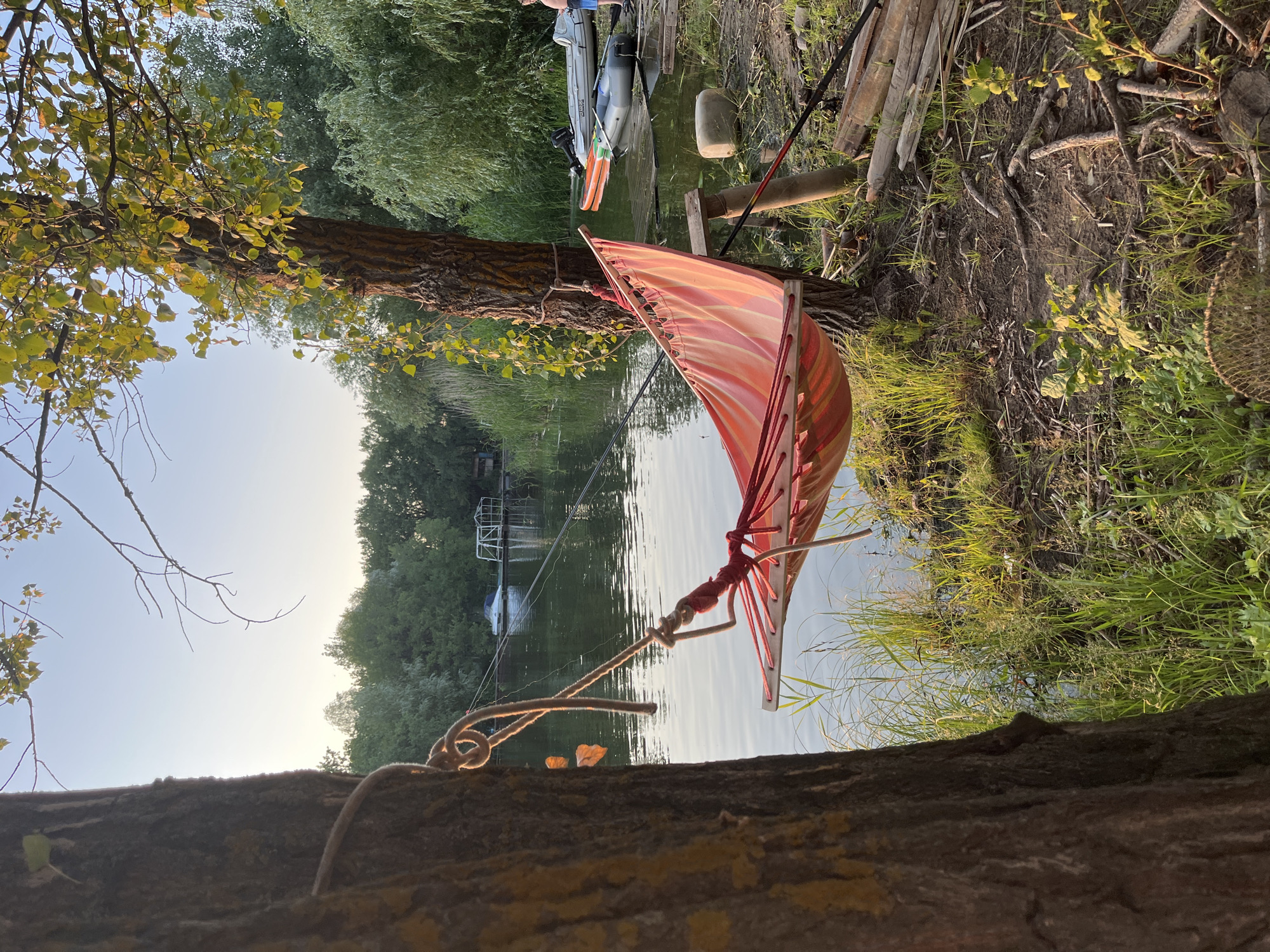 A brightly colored hammock, striped with pink and orange, is strung between two trees on the bank of a body of water, likely a river. The water is calm and reflects the sky, which is a pale blue with a hint of sunset warmth. Lush green trees and reeds line the opposite bank, creating a serene, natural backdrop. Several small boats are moored or resting on the water near the bank, including an inflatable boat with an outboard motor. On the shore, fishing rods are propped up, suggesting recreational activity. The ground is covered with dirt, grass, and fallen branches. A person, partially visible on the right side of the frame, appears to be standing by the water. The scene evokes a sense of relaxation and outdoor leisure. Given the context of Saratov, Russia, this location could be along the Volga River, a prominent feature of the city.