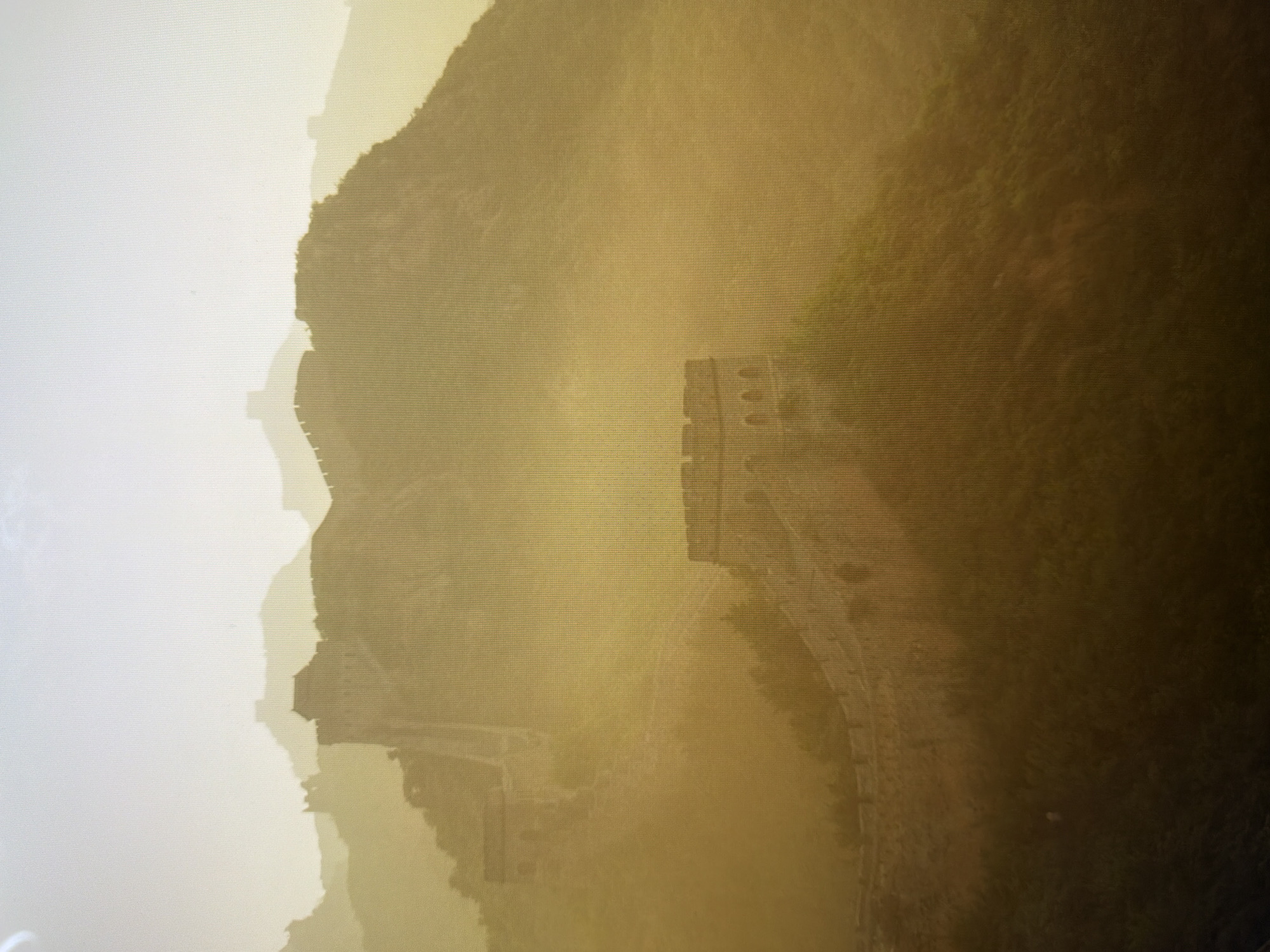 The image depicts the Great Wall of China stretching across a mountainous landscape under a hazy, golden light, suggesting either sunrise or sunset. The wall is a prominent stone structure with watchtowers, winding along the contours of the hills. Vegetation, likely trees and shrubs, can be seen sparsely growing on the slopes and along sections of the wall. The atmosphere is misty and diffused, softening the details of the distant peaks and the wall segments further away. No people or specific actions are visible. The scene is devoid of any elements relating to the city of Kyiv, Ukraine.