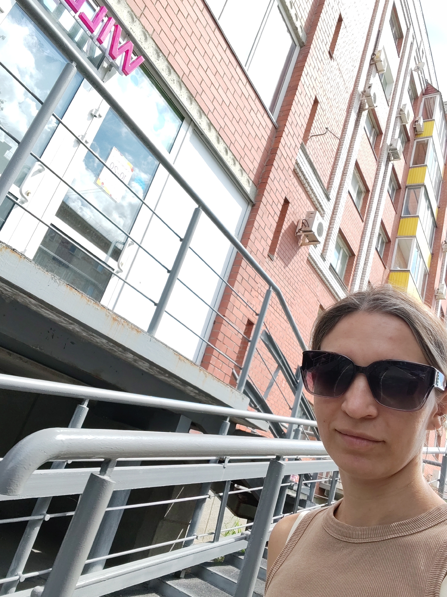 A woman with blonde hair and sunglasses is taking a selfie in front of a brick building. She is wearing a tan, ribbed tank top. The building has multiple windows, some of which reflect the sky. There is a metal railing in the foreground, and a sign with pink lettering that is partially visible on the building. The overall setting appears to be an urban environment.