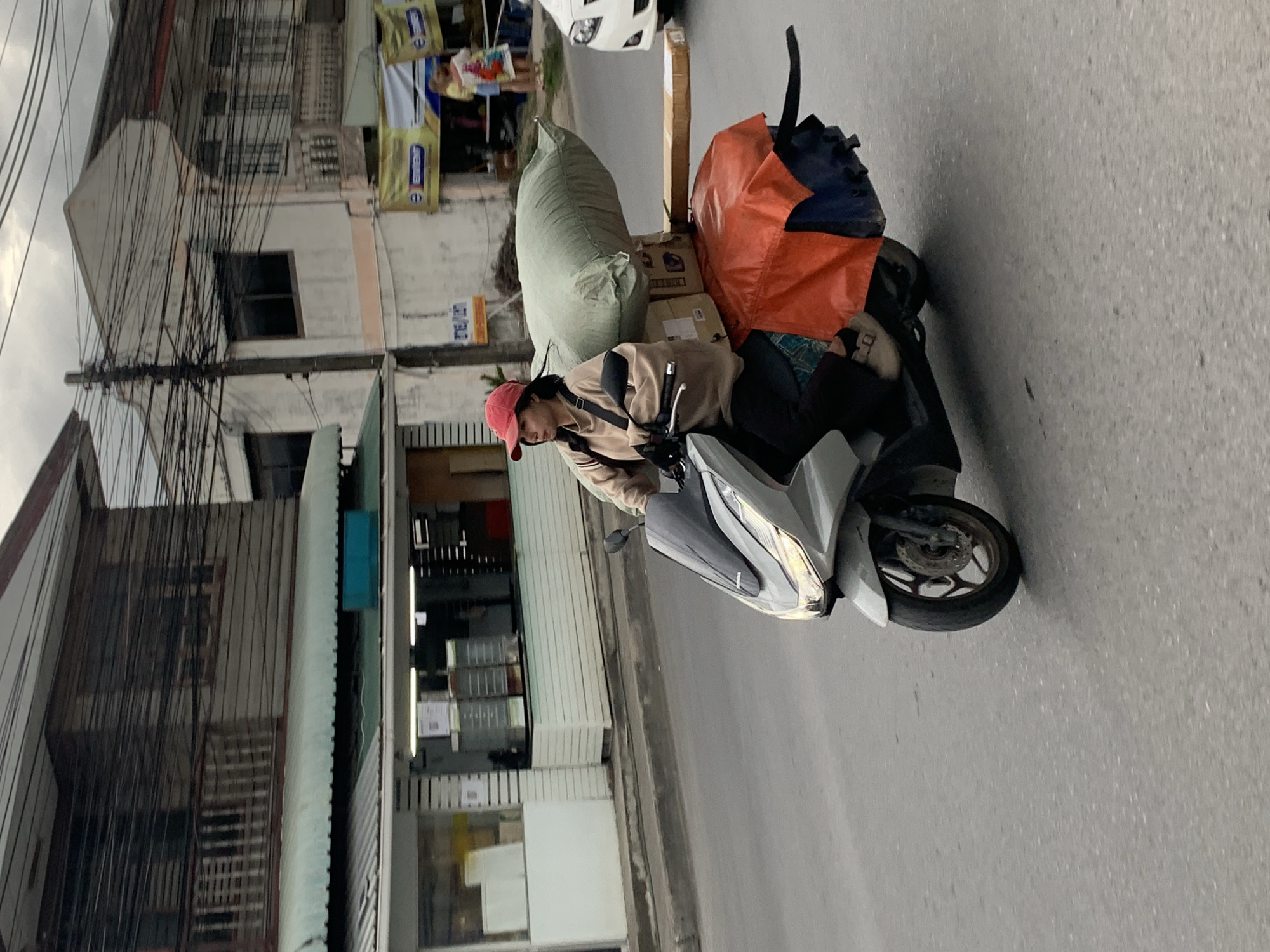 A woman wearing a red cap and a beige sweatshirt rides a silver scooter on a paved road in Ko Samui District, Thailand. She is carrying a large green bag and several boxes strapped to the back of the scooter, which is covered with an orange tarp. The scooter is in motion, as indicated by the blurred background. The surroundings show buildings with wooden and concrete facades, power lines, and some street signs. The sky is overcast.