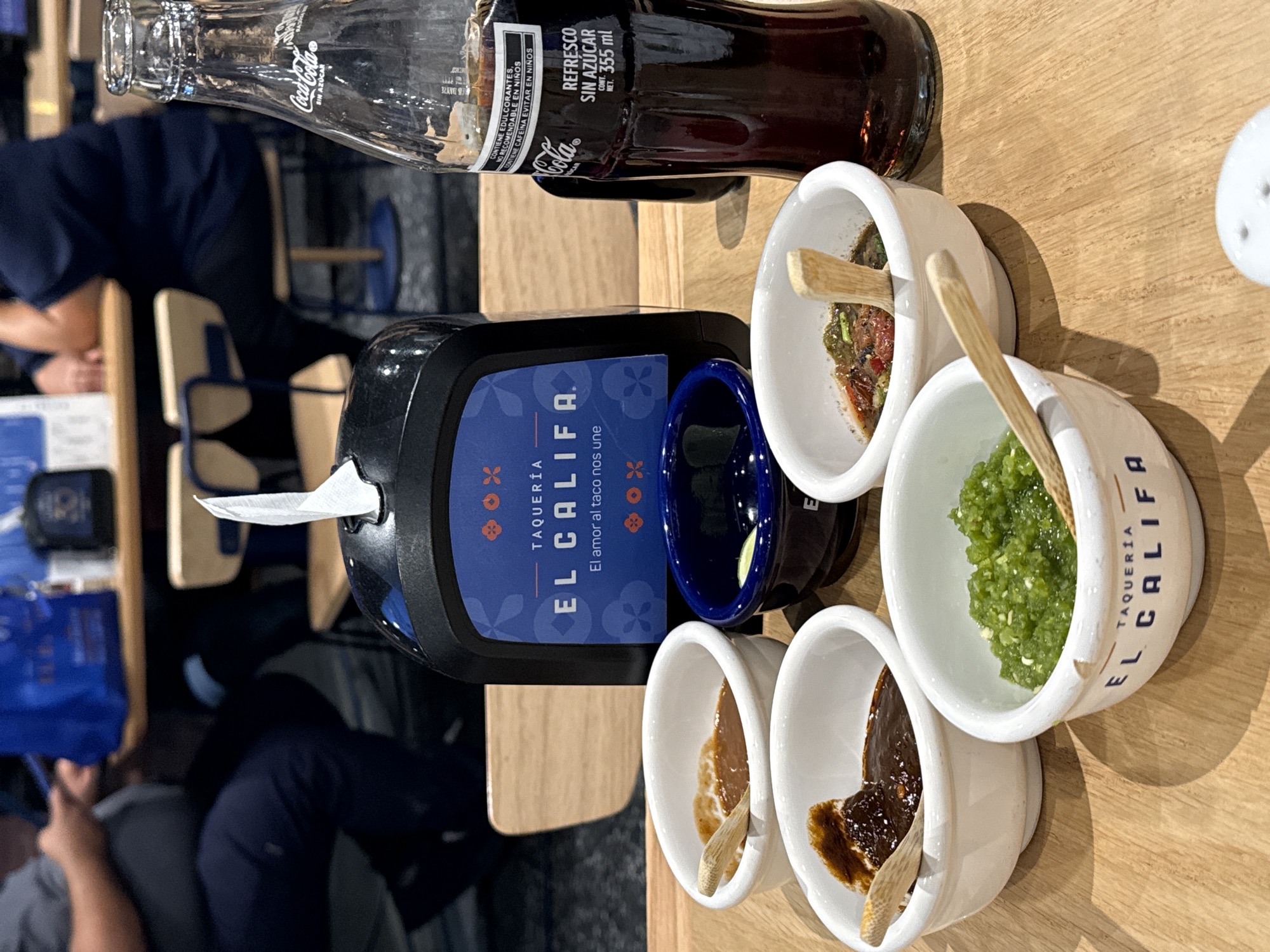 The image shows a table setting at "El Califa" taqueria, a restaurant chain in Mexico, specializing in tacos. The scene includes several small, white ceramic bowls containing various types of salsa and sauces, each with a wooden spoon. There's a dark blue bowl next to a black tissue dispenser with the restaurant's logo visible. A bottle of Coca-Cola Sin Azúcar (sugar-free) is also present. Part of a person wearing a dark blue shirt is visible. The table is light wood. The environment appears to be inside the restaurant. The city is not explicitly mentioned, but based on the OCR text, the restaurant is identified as "El Califa."