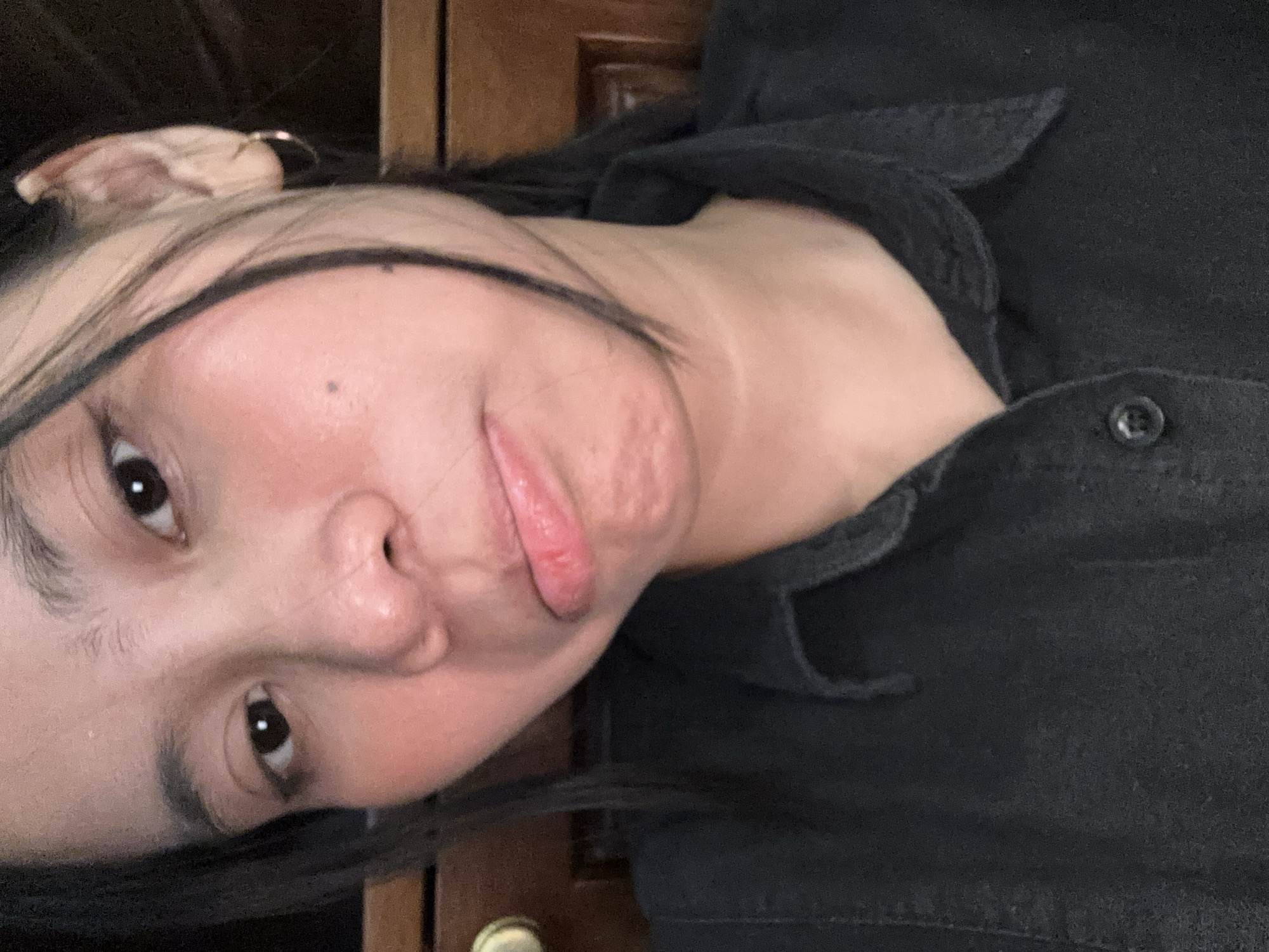 A close-up selfie of a young woman's face shows her making a pouty expression. She has dark hair with blonde highlights framing her face and is wearing a black collared shirt. Her right ear is visible and adorned with a small hoop earring. The background appears to be a wooden dresser. The image was taken in South Jakarta, Indonesia.