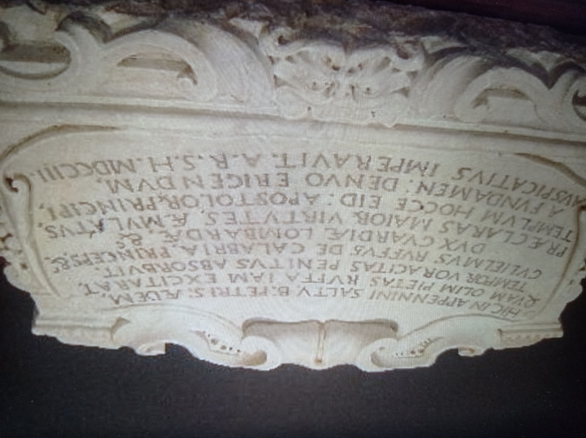 The image shows a close-up of a stone inscription. The inscription is in Latin and is carved into a decorative stone plaque. The text is arranged in several lines and appears to be an epitaph or memorial. The stone has ornate carved elements on the top and bottom. The lighting is somewhat dim, casting shadows that make some of the inscription difficult to read. The background is dark and out of focus. This inscription is located in Mukachevo, Ukraine.