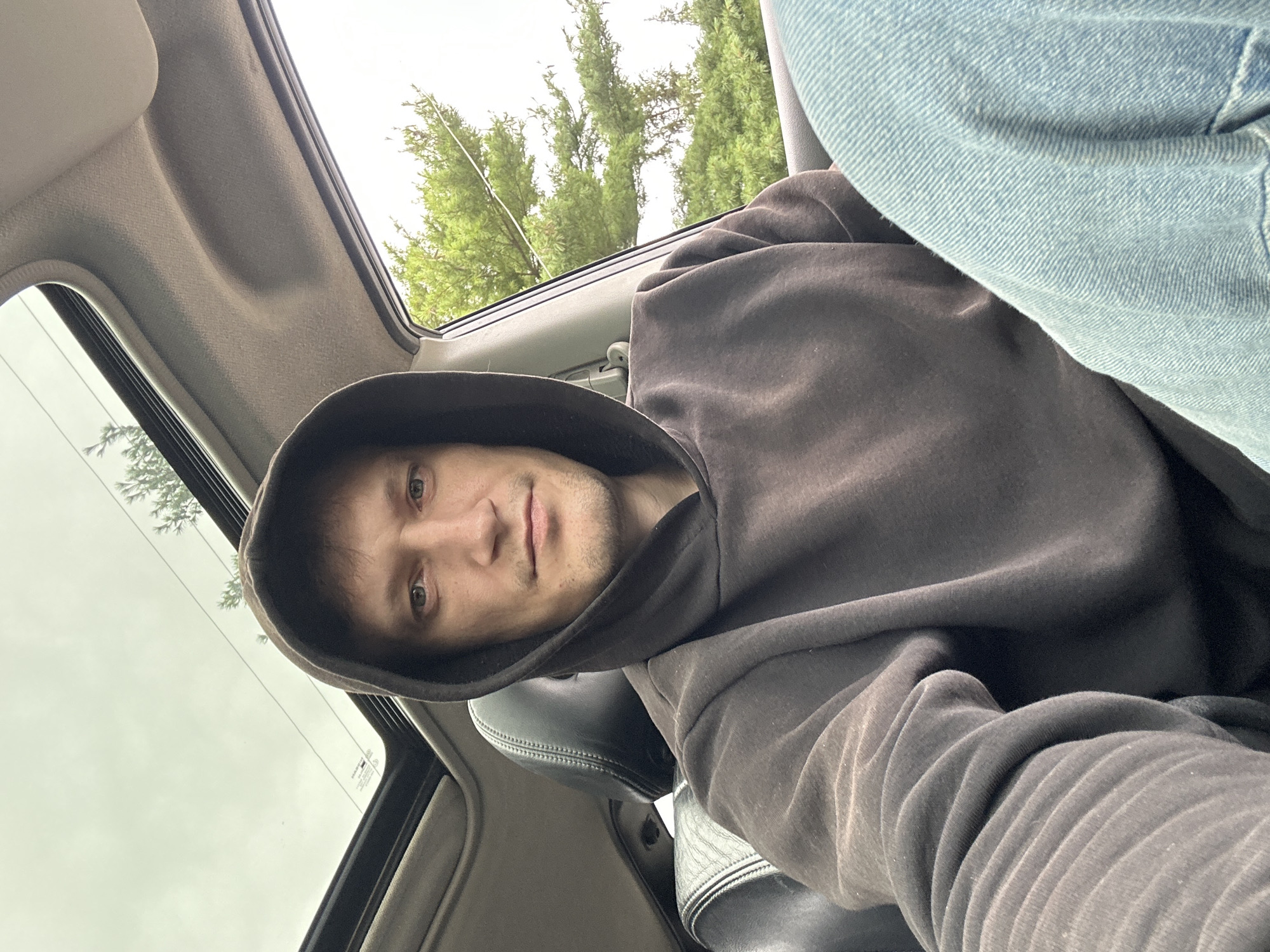 A man with light skin and short brown hair is sitting in the driver's seat of a car, looking directly at the camera. He is wearing a dark gray hooded sweatshirt and light blue jeans. The car's sunroof is open, revealing a cloudy sky and the branches of a green tree. The interior of the car is visible, including the black leather driver's seat and the gray headliner. The image appears to be a selfie taken from inside the vehicle. No specific location within Makiv, Ukraine is identifiable from the image.