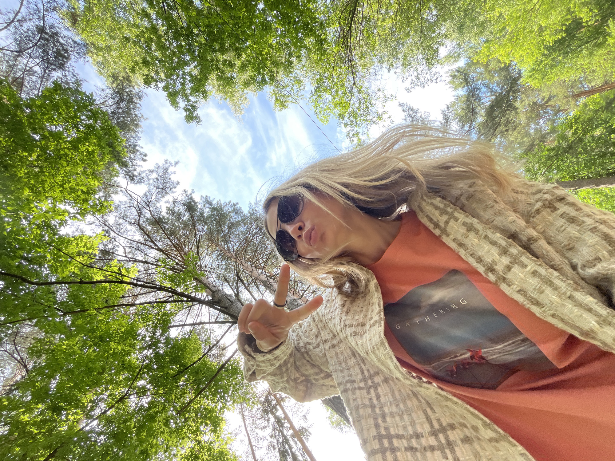A low-angle selfie captures a young woman looking up into a vibrant forest canopy and sky in Chaikovsky, Russia. The blonde-haired woman, wearing sunglasses, an orange t-shirt, and a light beige patterned jacket, poses playfully for the camera. She makes a peace sign with her right hand, which has a black ring on her index finger, and pouts her lips as if blowing a kiss. Her hair appears to be flowing slightly to her right.
The background consists of a bright blue sky with scattered white clouds, framed by the lush green foliage of various trees, including both deciduous trees with broad, bright green leaves and coniferous trees with darker needles. Sunlight filters through the leaves, creating dappled light and highlighting the vibrant greenery. The low perspective emphasizes the height of the trees and the vastness of the sky. The word "GATHERING" is visible in white text on the front of her orange t-shirt, above a graphic that appears to show a dark, wave-like scene with hints of red. The overall mood is casual and cheerful, set on a bright, clear day outdoors.