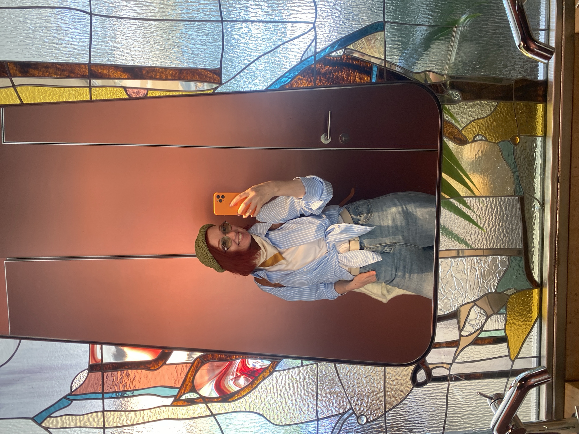 The image, taken in Sochi, Russia, shows a mirror selfie of a young adult female in an indoor setting. The central subject is the woman, who is reflected in a large, tall mirror with a dark reddish-brown frame. She has reddish-brown hair and is wearing a green knit beanie, round sunglasses, a white undershirt, a light blue and white striped button-up shirt tied at the waist, and blue jeans. She is holding an orange smartphone, likely an iPhone, up to capture her reflection in the mirror, and is smiling broadly at the camera.
Behind and around the mirror is a vibrant stained-glass window, which serves as a prominent background element. The window features various textured and colored glass panels, including clear, light blue, yellow, amber, brown, and some sections with swirling red and orange patterns. These panels create an abstract design, allowing natural light to filter into the space and cast colorful reflections. A door handle and a lock mechanism are visible on the brown surface of what appears to be a door or wall section next to the mirror. Parts of a green plant are also faintly visible, reflected in the mirror and through the stained glass. The overall scene suggests a brightly lit interior, possibly an entryway or hallway, with decorative elements. There is no visible text in the image.