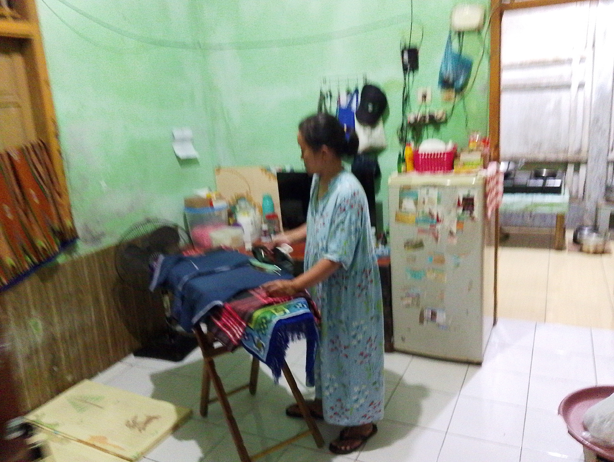 The image depicts an indoor scene, likely a room in a residential dwelling within Desa Salebu Kecamatan Majenang, Indonesia. A woman, appearing to be of adult age, is standing and engaged in an activity involving a wooden folding stand on which several textiles are draped, including what appears to be folded blue fabric and a patterned cloth with tassels. The woman is wearing a long, light blue patterned dress. To the right of the woman is a white refrigerator adorned with various magnets and papers. Behind the woman, a green wall is visible, with items such as bags and electrical fixtures mounted on it. To the left of the woman, a wooden door frame and a portion of a colorful textile are visible. The floor is tiled in a white grid pattern. A black fan is positioned behind the wooden stand. The overall impression is of a domestic setting where laundry or textile work is taking place.