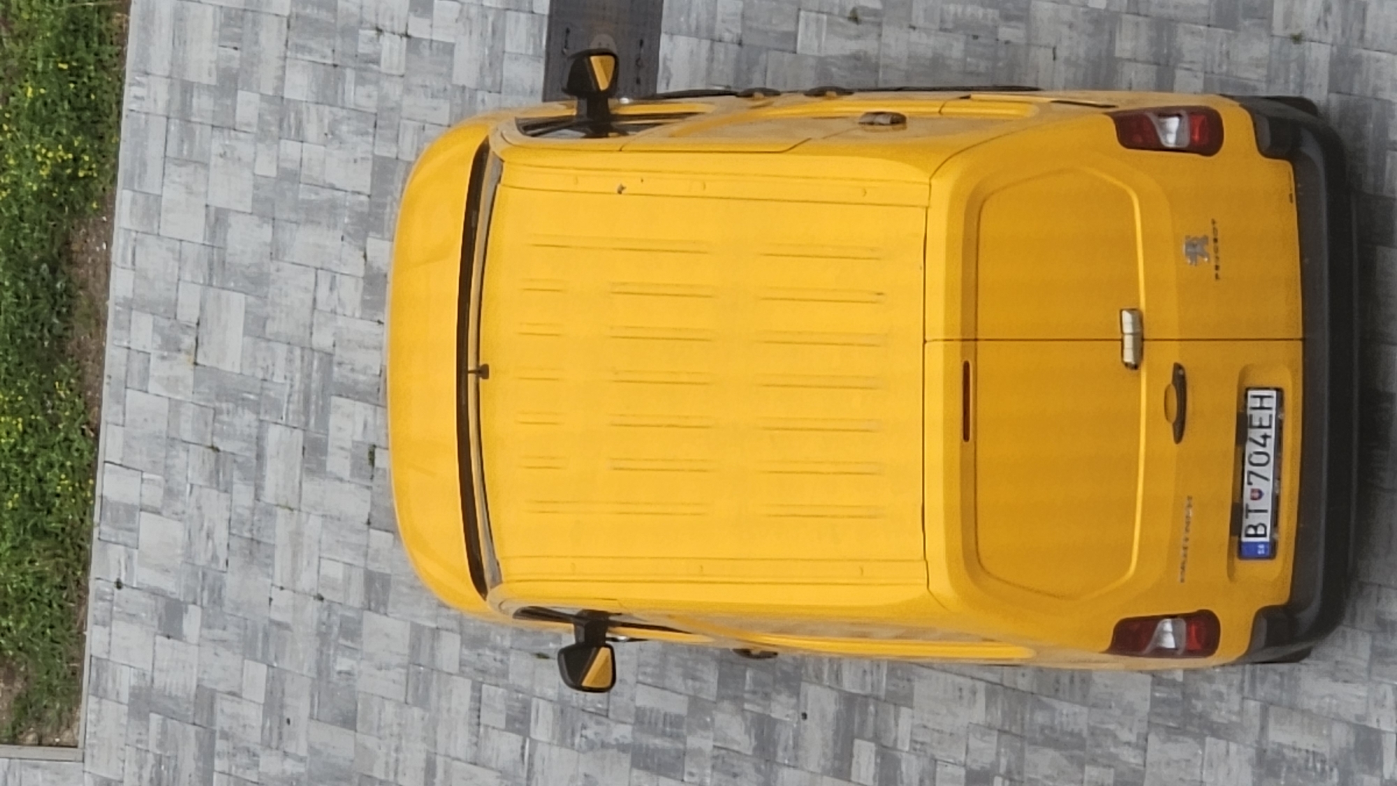 An aerial, top-down shot shows the rear of a bright yellow Peugeot van parked on a patterned grey stone pavement. The van's rear doors are closed, and a license plate reading "BT 704EH" is visible. The van is positioned in what appears to be an outdoor urban setting, possibly a parking area or a street, adjacent to a strip of green grass with small yellow flowers. The pavement is made of rectangular grey stones laid in a repeating pattern. The lighting suggests it is daytime.