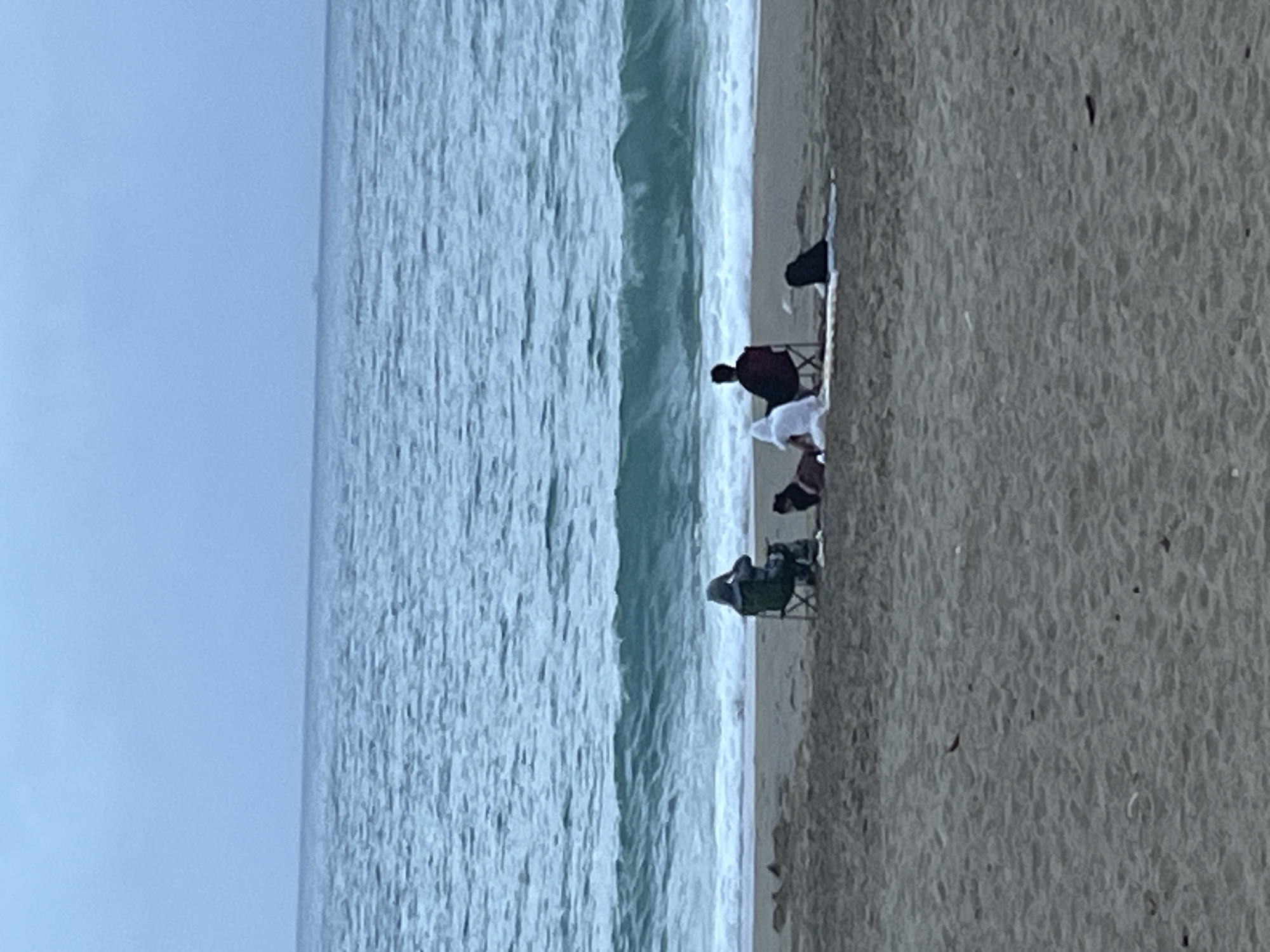 The image shows a beach scene in Alanya, Turkey. Several people are sitting on the sand near the water's edge. Some are seated in chairs, while others are sitting directly on the ground. There is a visible shoreline, ocean waves, and sky. The overall scene suggests a calm and casual gathering.