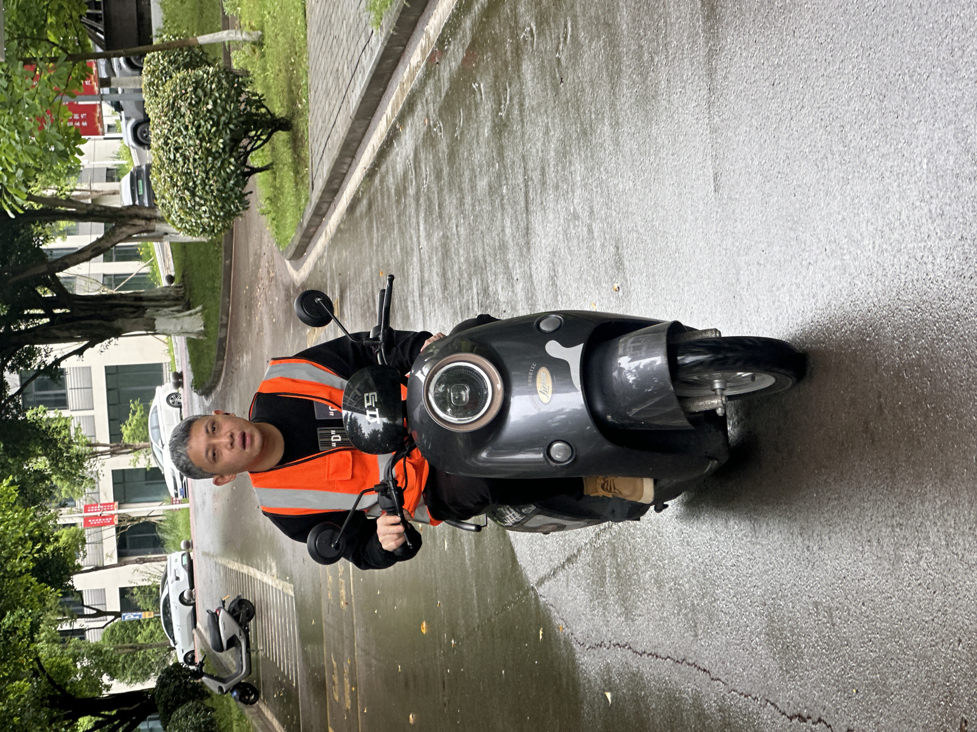 The image displays a man seated on a dark grey electric scooter on a wet asphalt path or road. The man, with short grey hair, is wearing a black long-sleeved shirt and a bright orange reflective safety vest adorned with white horizontal stripes. He looks directly at the camera with a neutral expression. Partial text, including "S'D" and "D"", is visible on his vest. The scooter features a prominent circular headlight with a light brown or gold trim, and the word "NERGETIC" is partially legible on its body.
The setting is an outdoor environment, characterized by green trees, manicured bushes, and multi-story buildings in the background, suggesting a park, campus, or residential area. The ground is visibly wet, indicating recent rainfall. A paved sidewalk or curb runs alongside the road. Further down the path, a white sedan, a grey SUV, and another white electric scooter are parked. A small wooden block is visible on the ground near the front wheel of the man's scooter. The scene is captured during daytime, likely under an overcast sky.