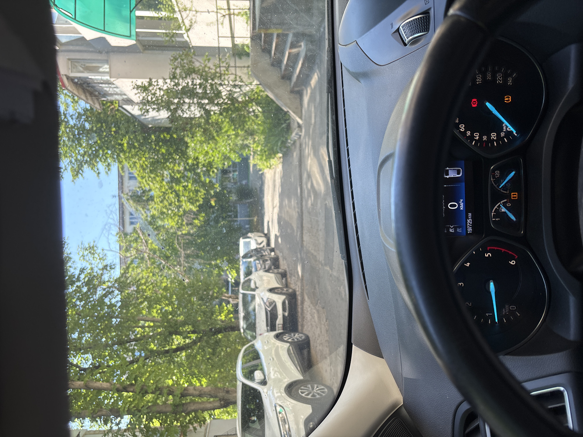 The image shows a view from inside a car, looking out through the windshield. The car is parked on a street lined with trees and apartment buildings. In the foreground, the dashboard and steering wheel of the car are visible. The dashboard displays various gauges and indicators, including a speedometer showing 0 km/h and an odometer reading 197725 km. Several other cars are parked on the street. The environment appears to be a residential area in Chișinău, Moldova, given the architecture of the buildings and the presence of a Moldovan license plate on one of the cars. The sky is blue and there are shadows cast by the trees, indicating it is daytime.