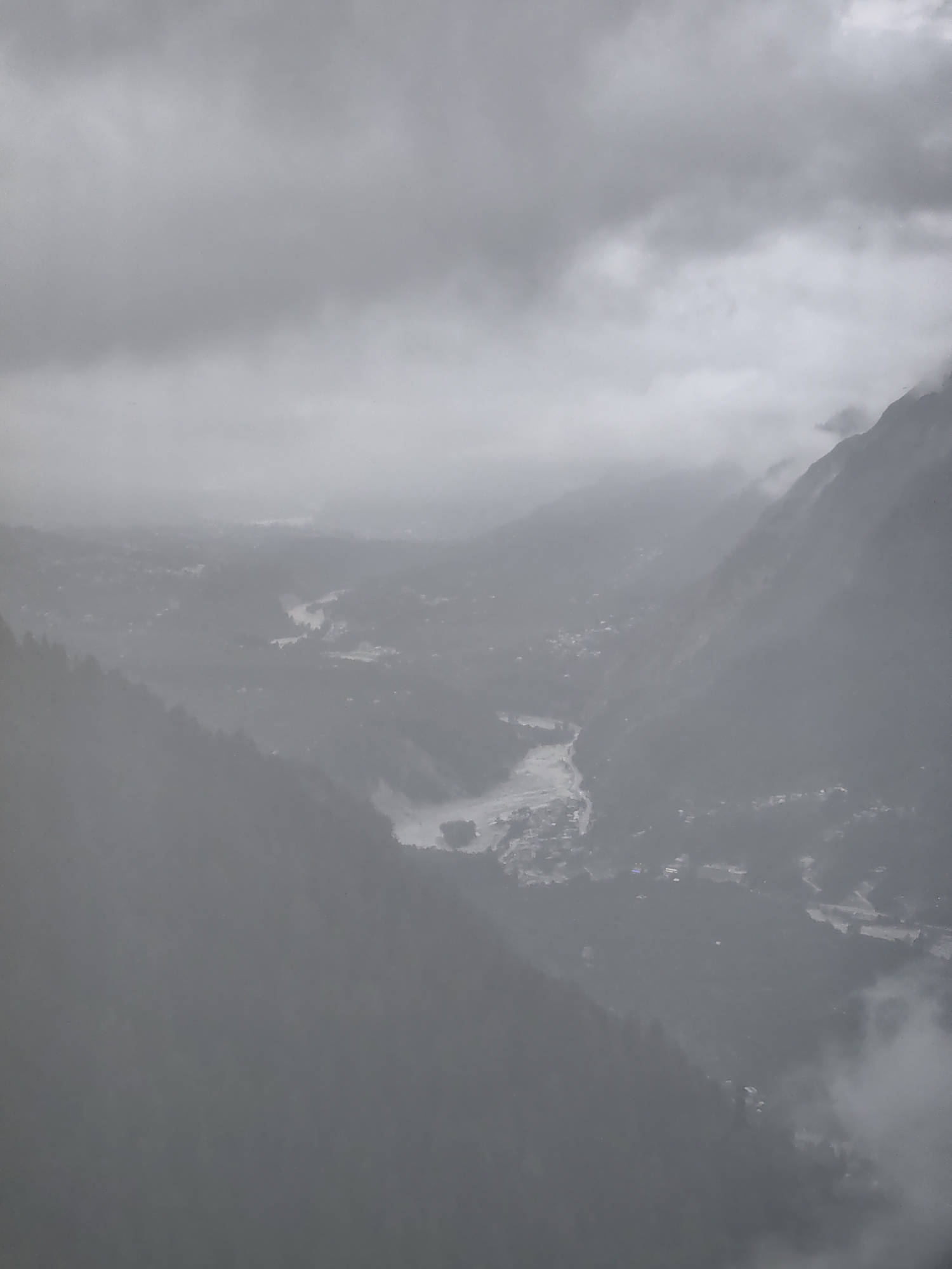 An aerial, high-angle, long shot depicts a hazy, overcast day in the mountainous region of Prini, India. A winding, muddy river cuts through a valley flanked by steep, tree-covered slopes. Scattered settlements, appearing as small clusters of buildings, are visible on the valley floor and hillsides. The sky is heavily clouded, obscuring any clear view of the sun or distant features. The overall atmosphere is one of subdued light and atmospheric obscurity.